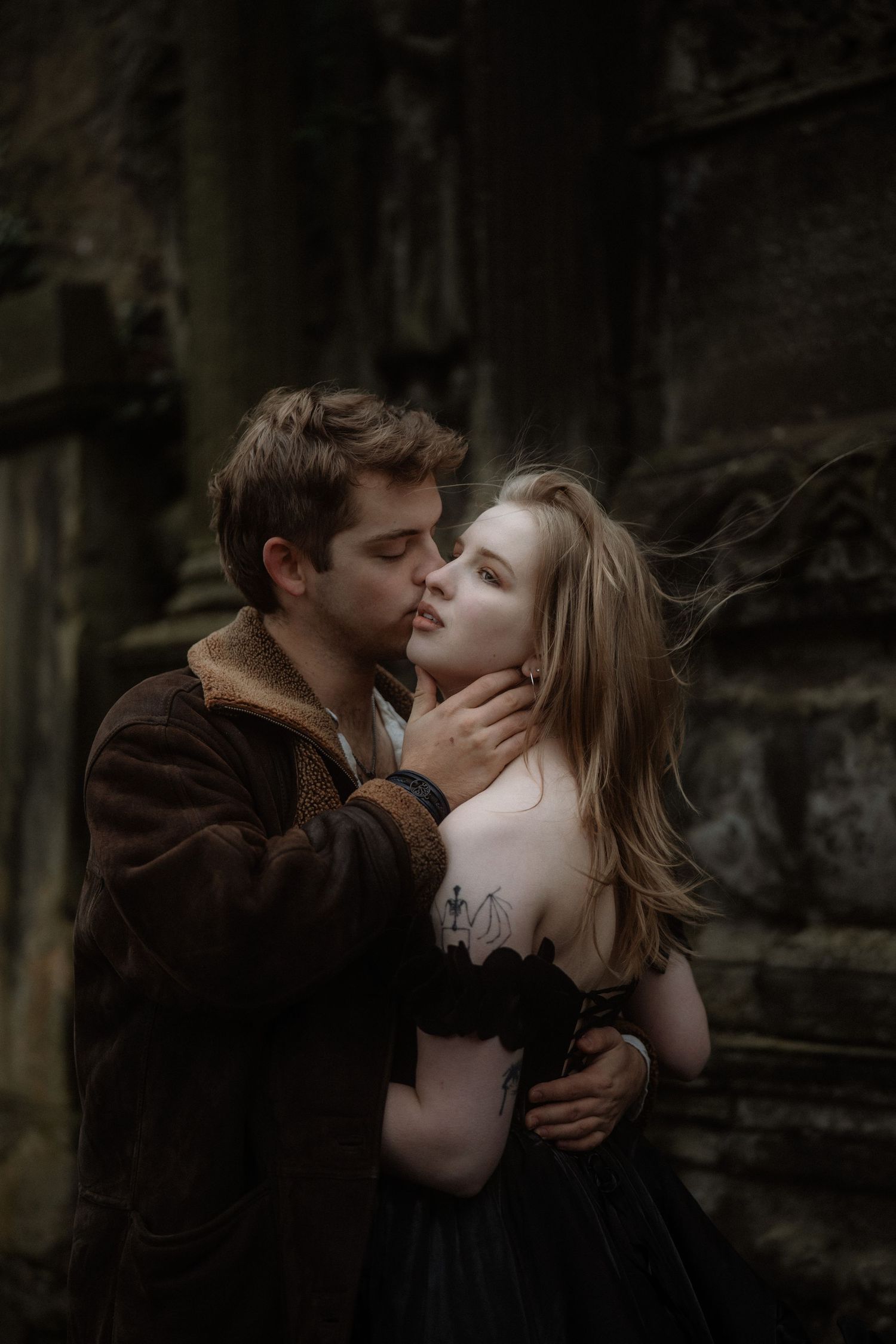 A romantic couple in dark gothic attire share an intimate moment against ancient stone ruins.