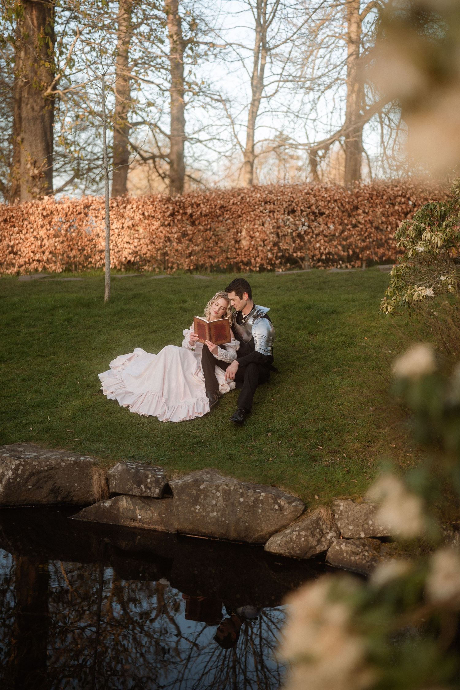 Bride and groom sitting together on a grassy hillside near a pond, surrounded by autumn trees in a romantic outdoor setting.
