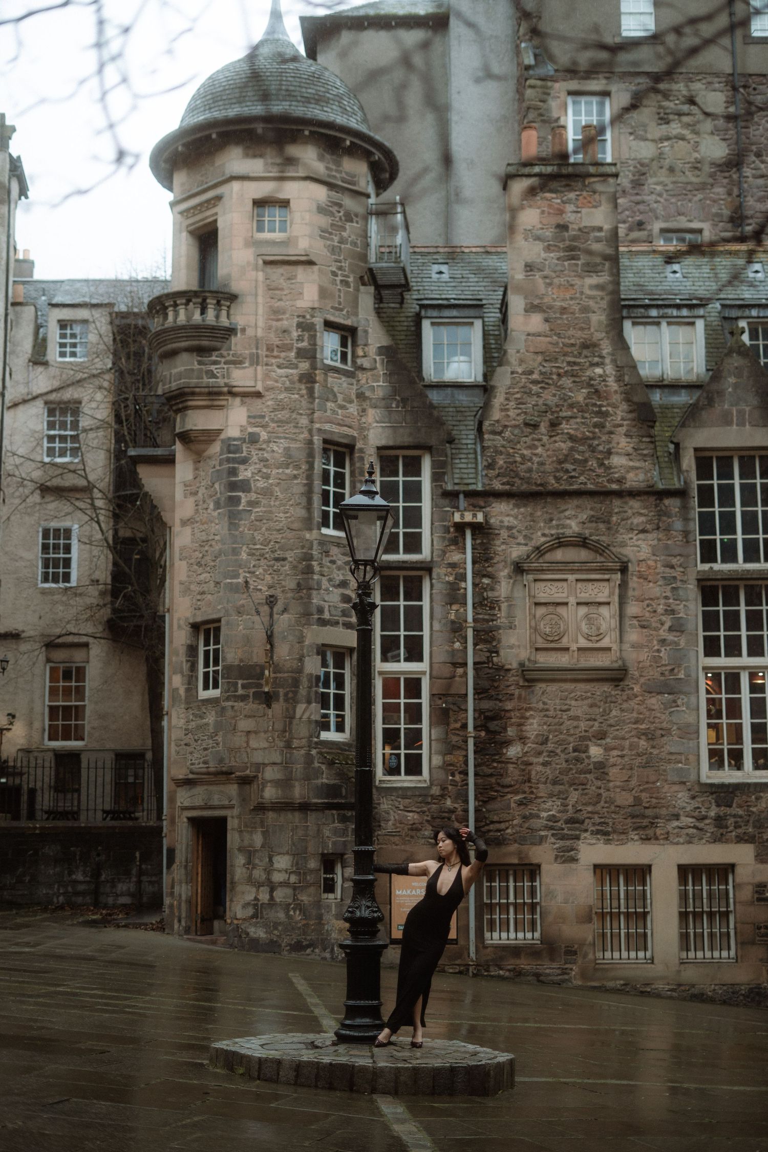 A figure in black stands against a historic stone building with turrets in Edinburgh's Old Town on a rainy day.