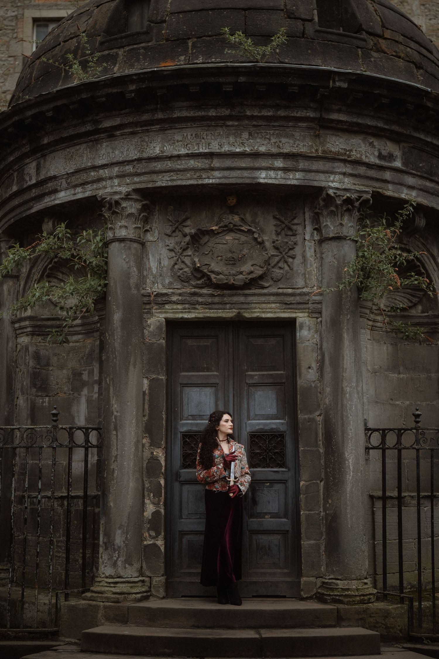 Woman in ornate jacket stands before ancient stone mausoleum with carved details and weathered wooden door.