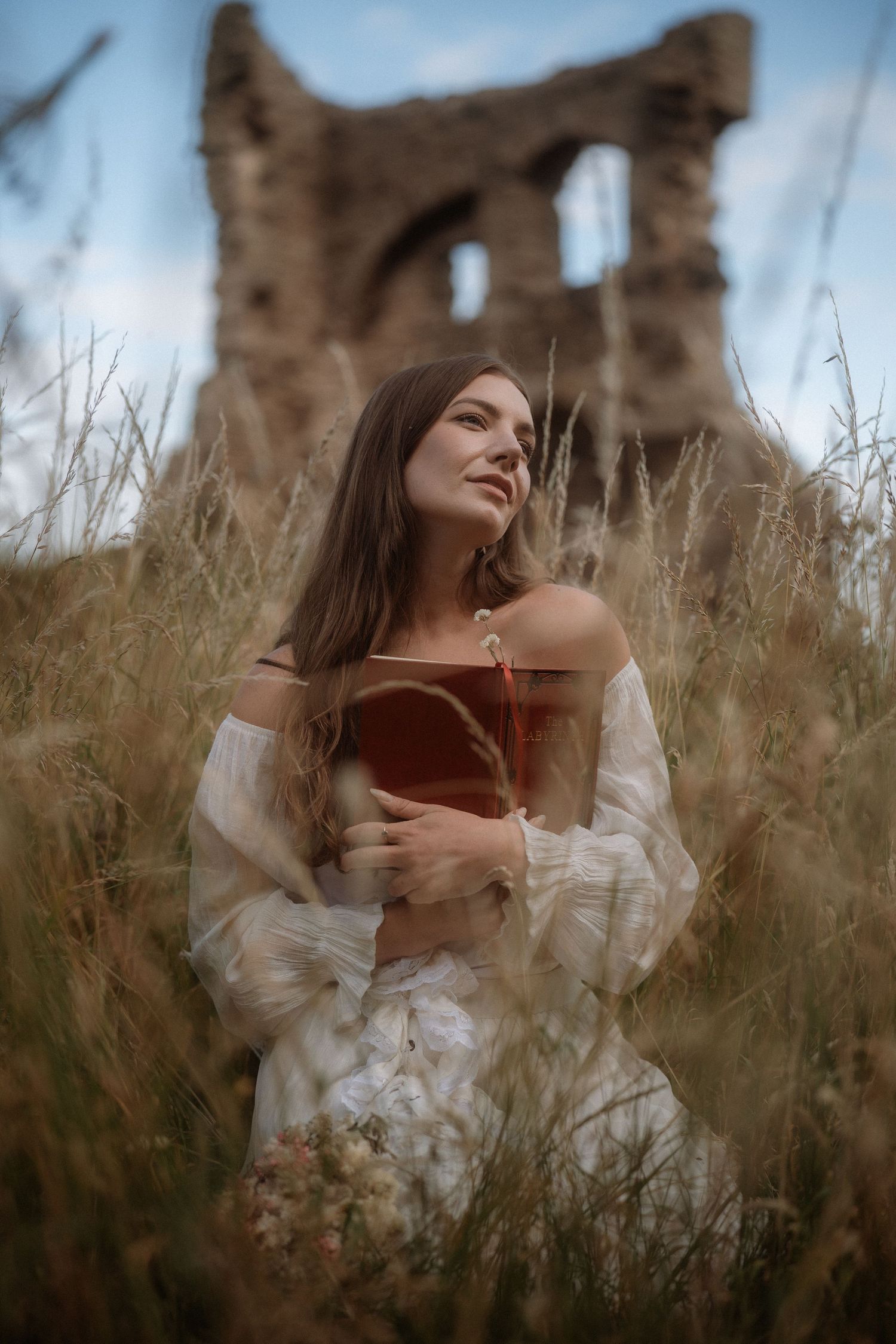 Young woman in white off-shoulder dress holds book in tall grass field with ancient stone ruins in background.