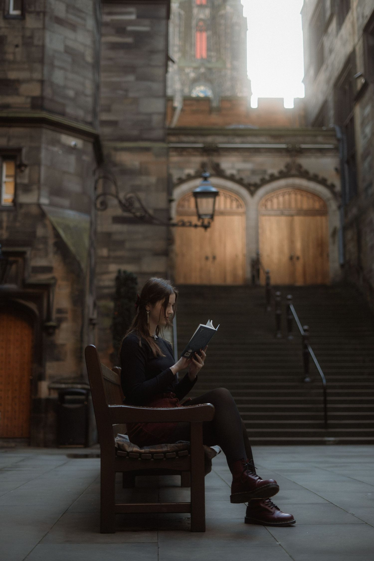A young woman sits in a wooden chair reading a book outside a historic stone building with ornate architecture.