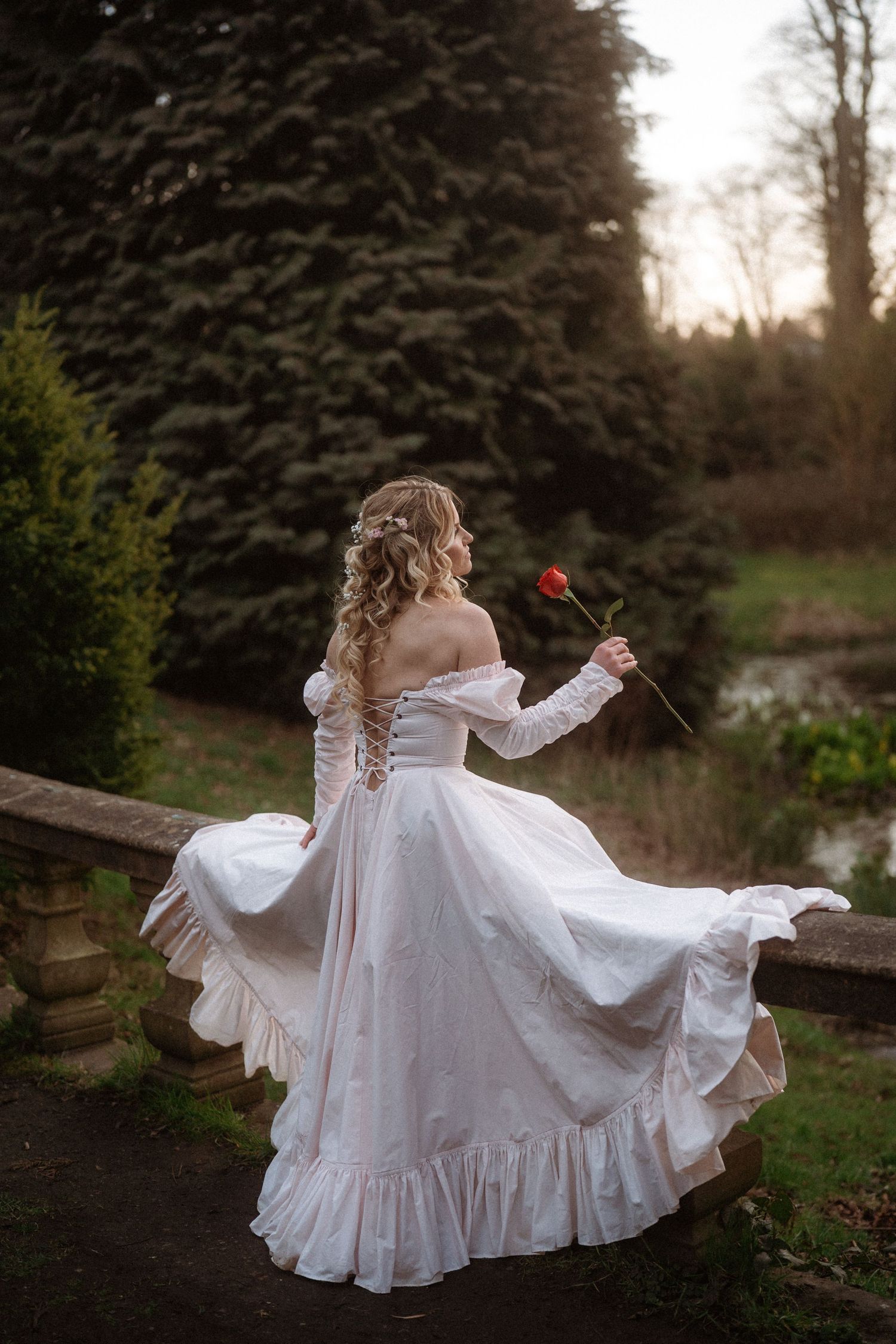 A woman in a flowing white off-shoulder gown holds a red rose in a garden at sunset.