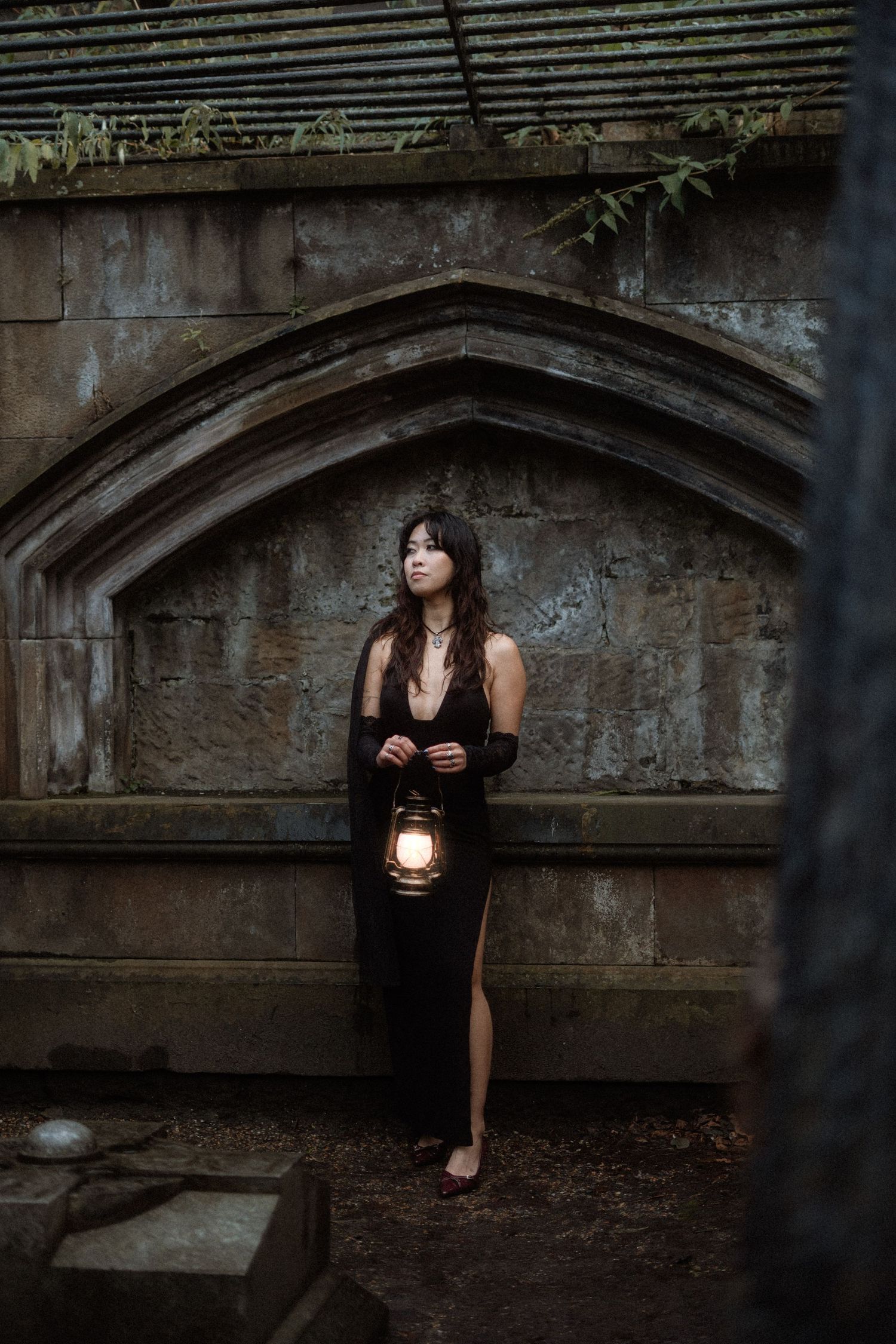 A dark and moody scene of someone in a black dress holding a lantern against an old stone archway background.