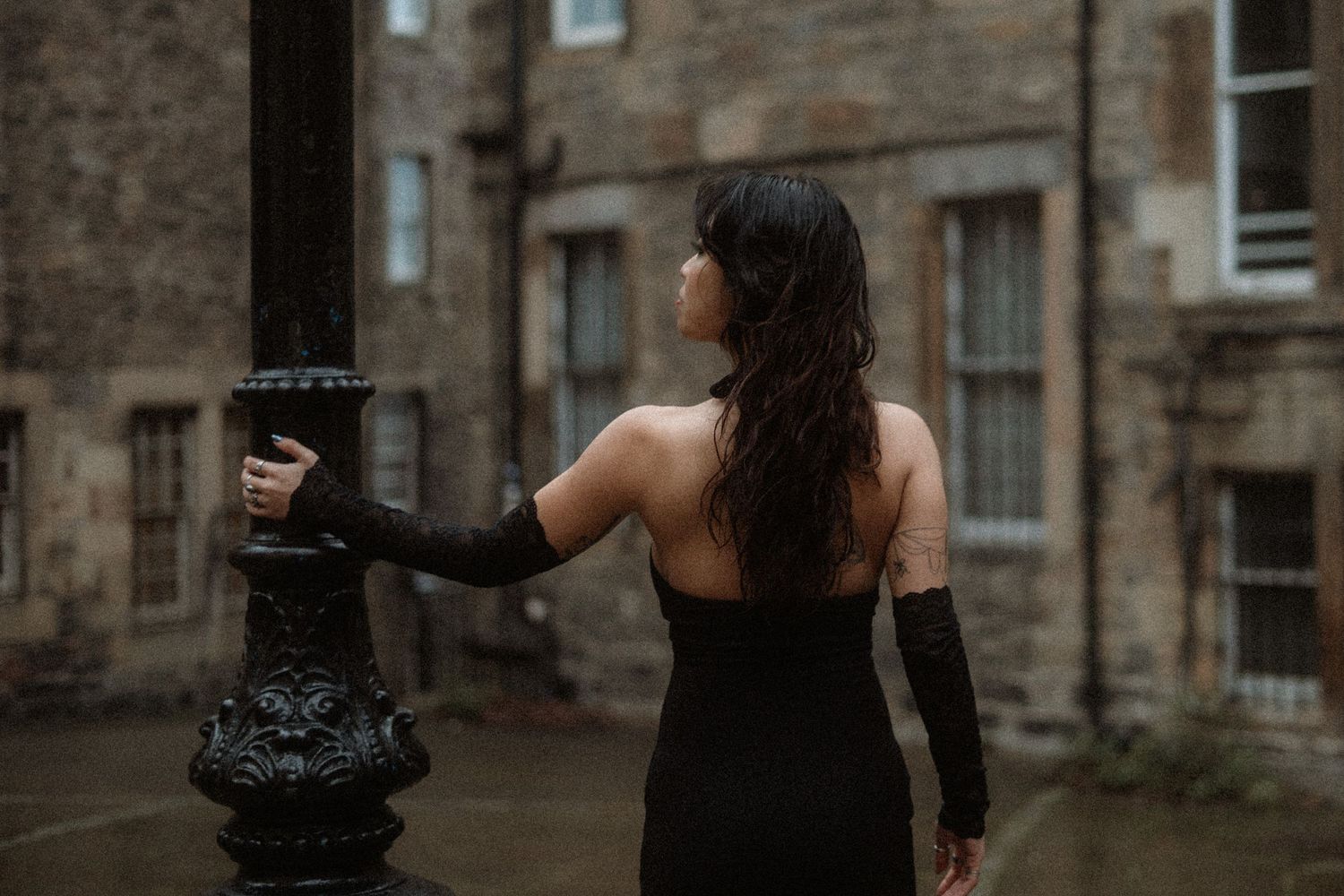 A woman in a black dress stands against a stone wall while reaching out her hand in a dramatic pose.