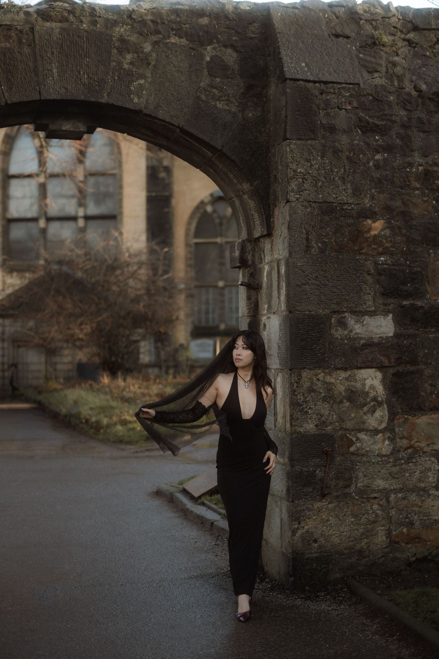 A person in a black dress poses gracefully against stone archways and walls in a moody outdoor setting.