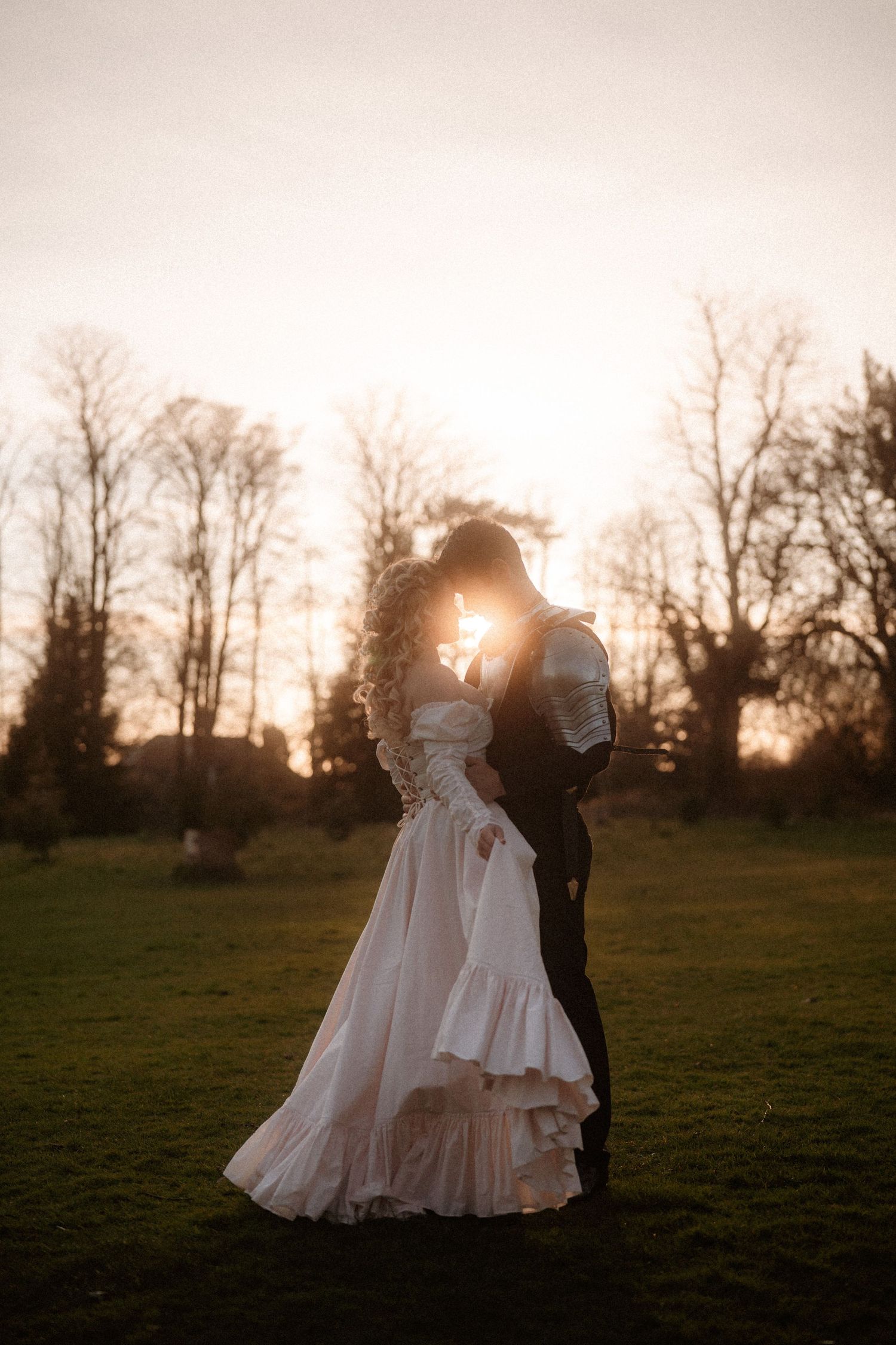 Bride and groom silhouetted against golden sunset light, sharing romantic moment in lush green field with bare winter trees.