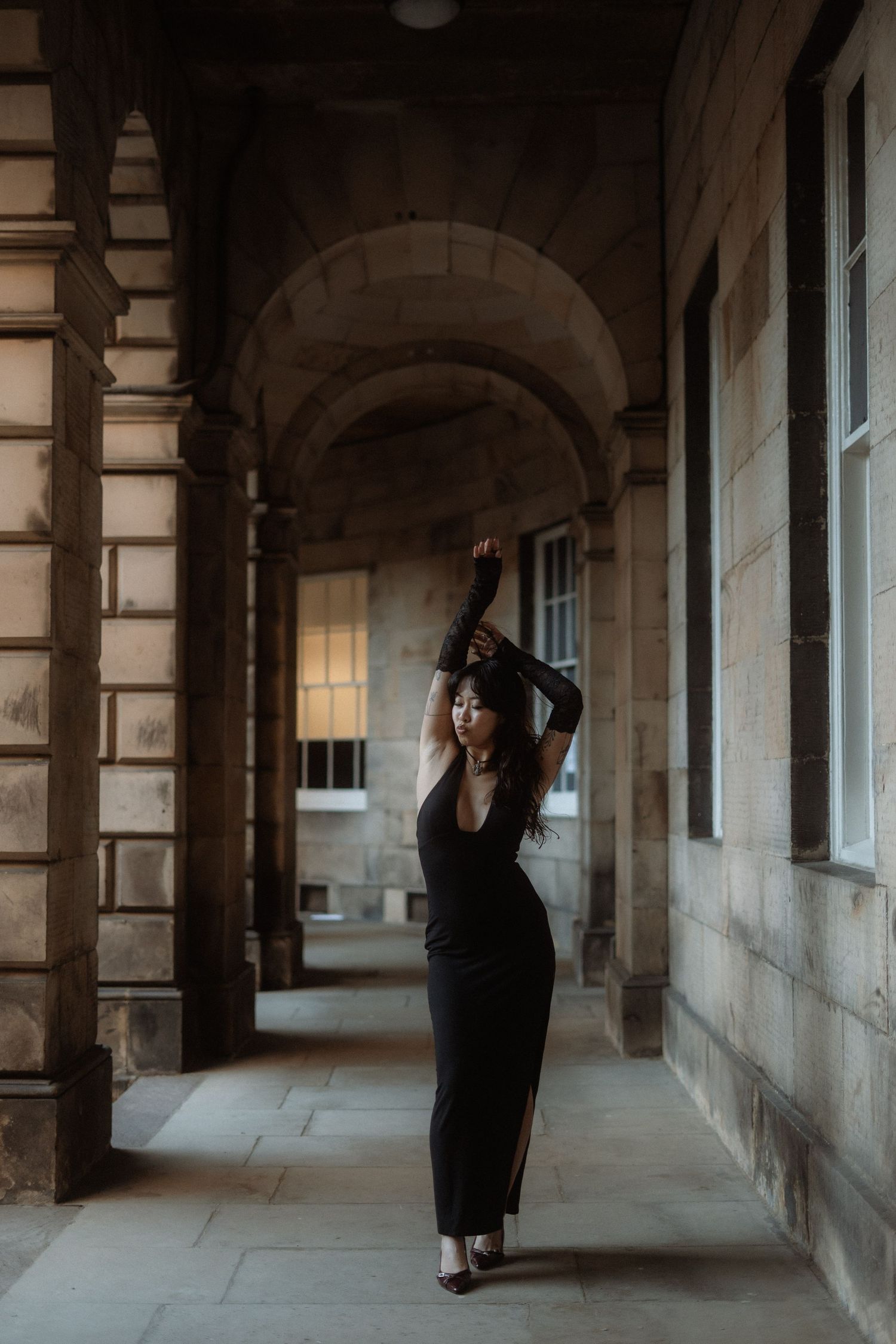 A dancer in a black dress strikes an elegant pose beneath stone archways in a historic corridor.