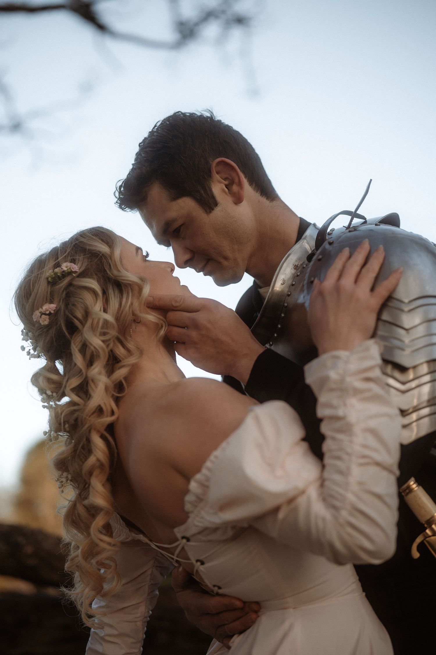A couple in medieval attire share a romantic moment, the man in armor tenderly holding the woman in a white gown.
