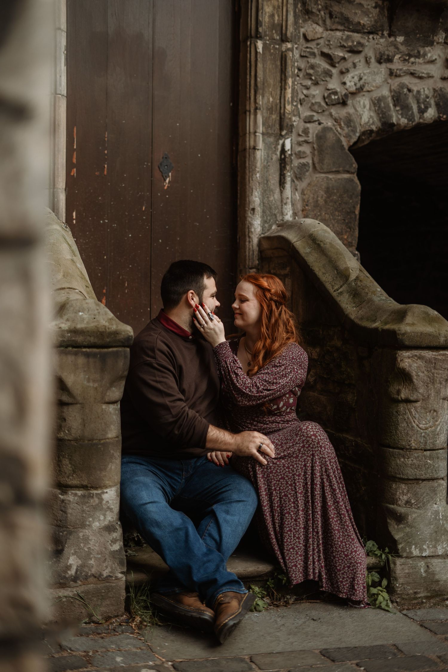 A couple shares a romantic moment on stone steps of an old building, woman in floral dress touching mans face.