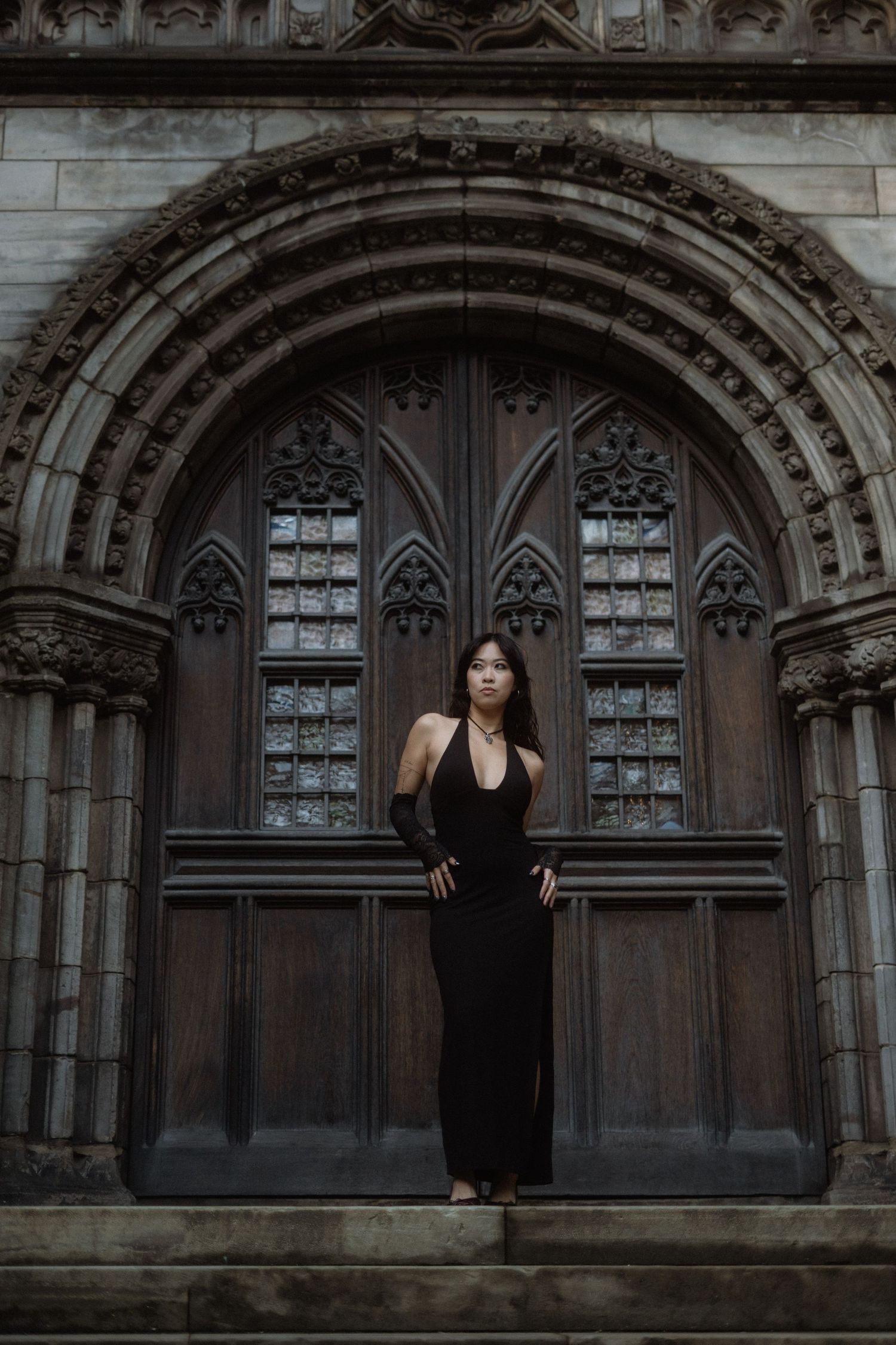 A figure in black dress poses dramatically against a gothic stone archway with ornate architectural details.