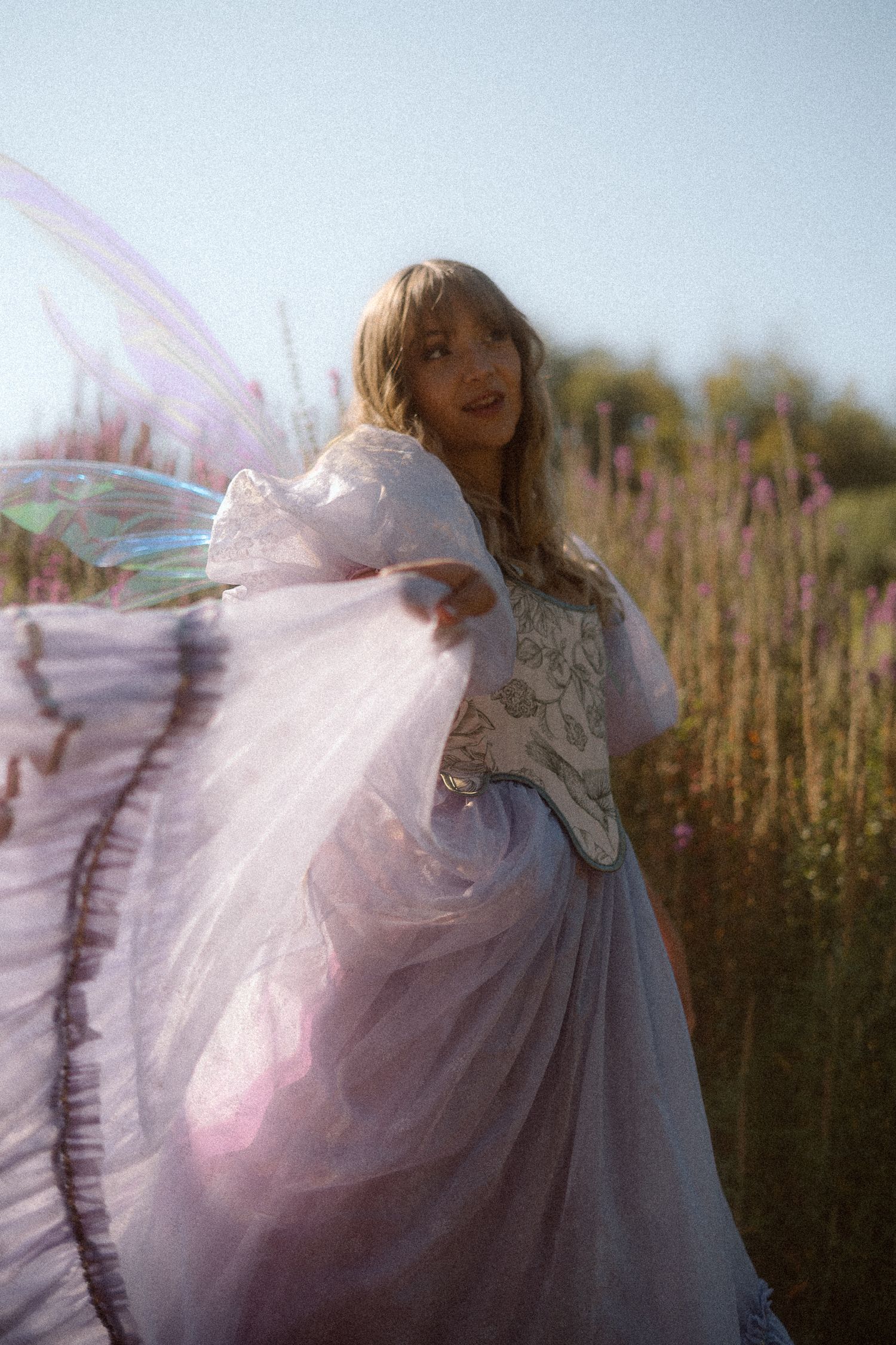 Ethereal figure in flowing white dress and fairy wings twirls in a sunlit meadow with tall grass and flowers.