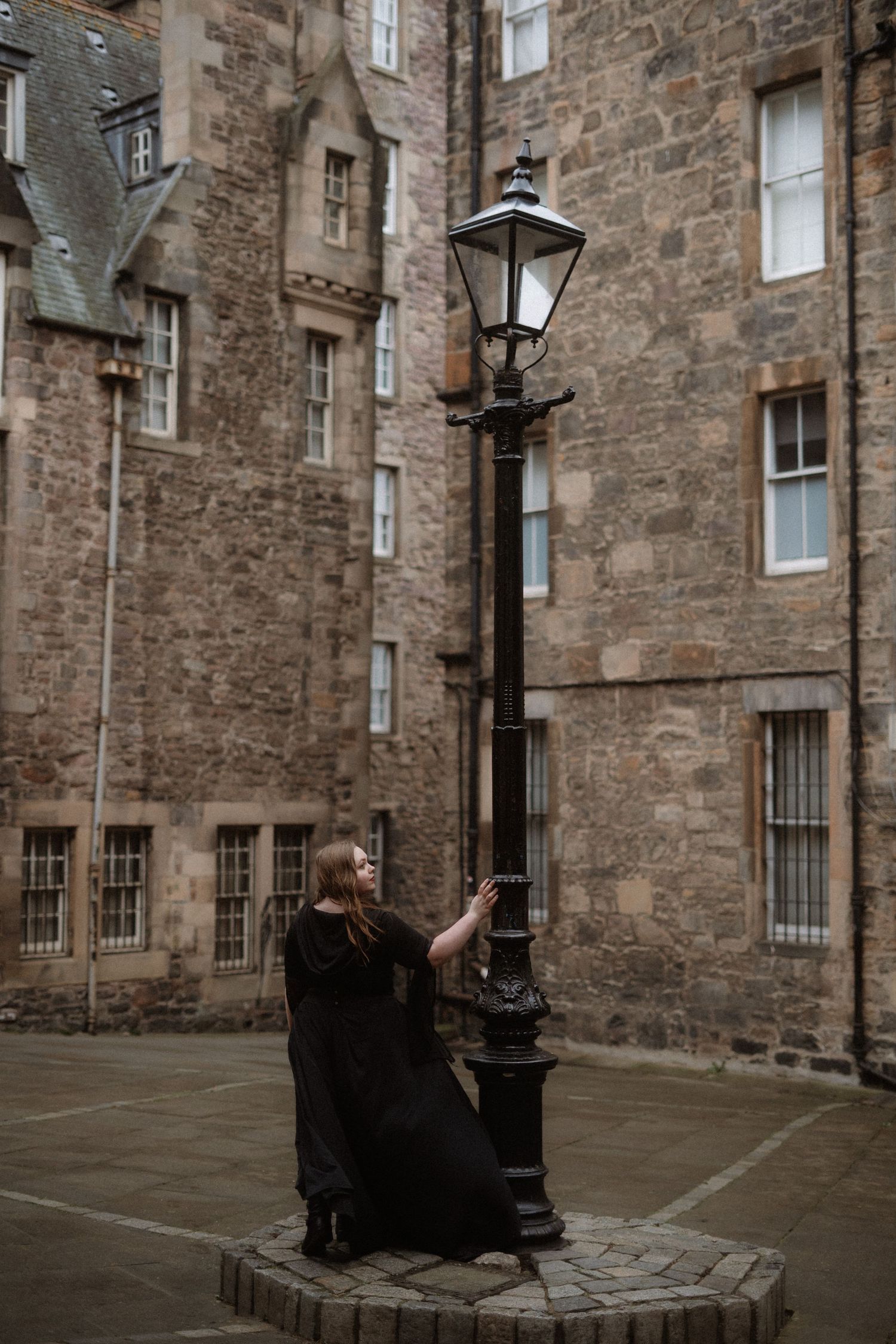 Figure in a black dress poses against a vintage lamp post in a historic stone courtyard in Edinburgh, Scotland.