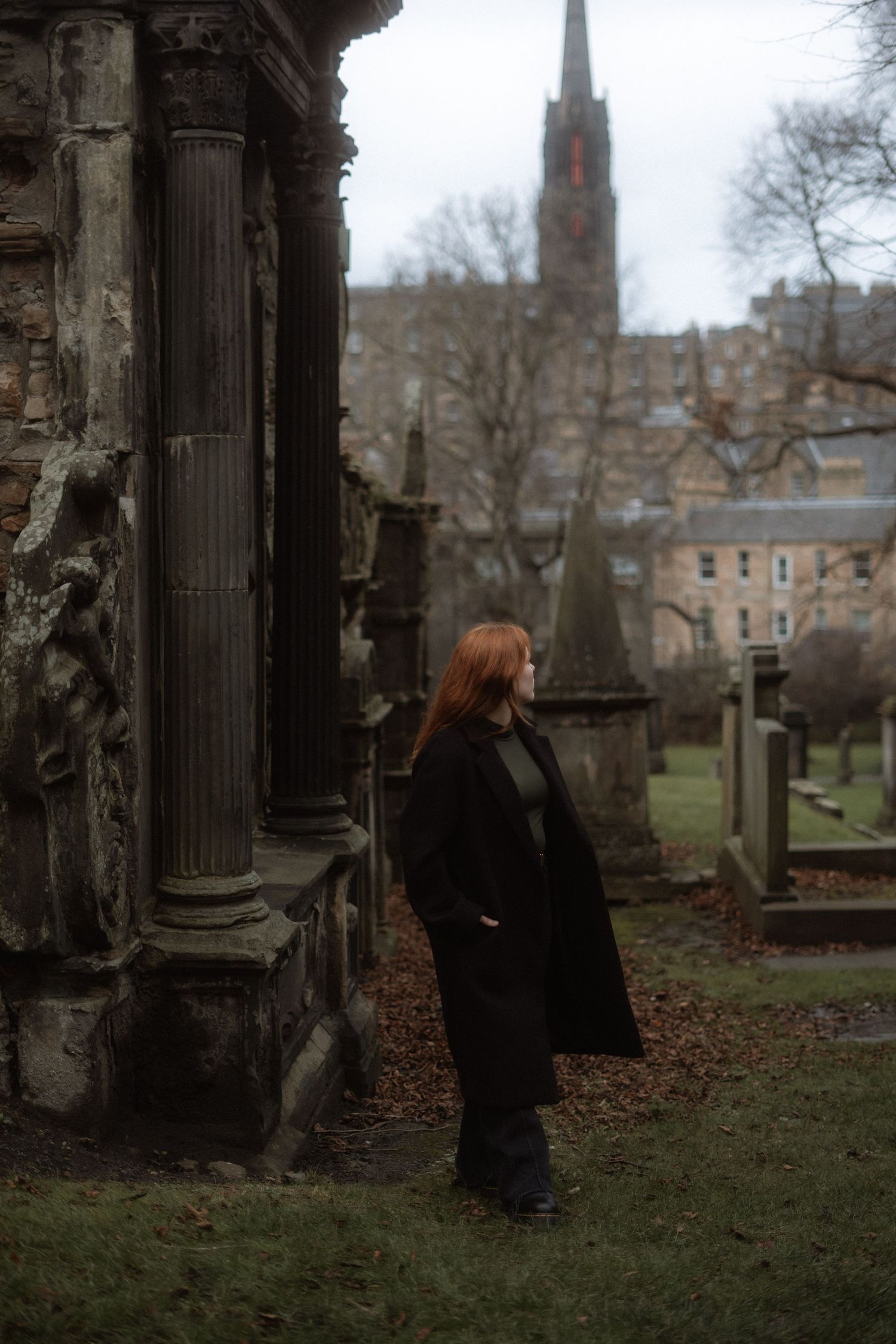 Red-haired woman in black coat standing beside ancient stone monument in historic cemetery with church spire in background.