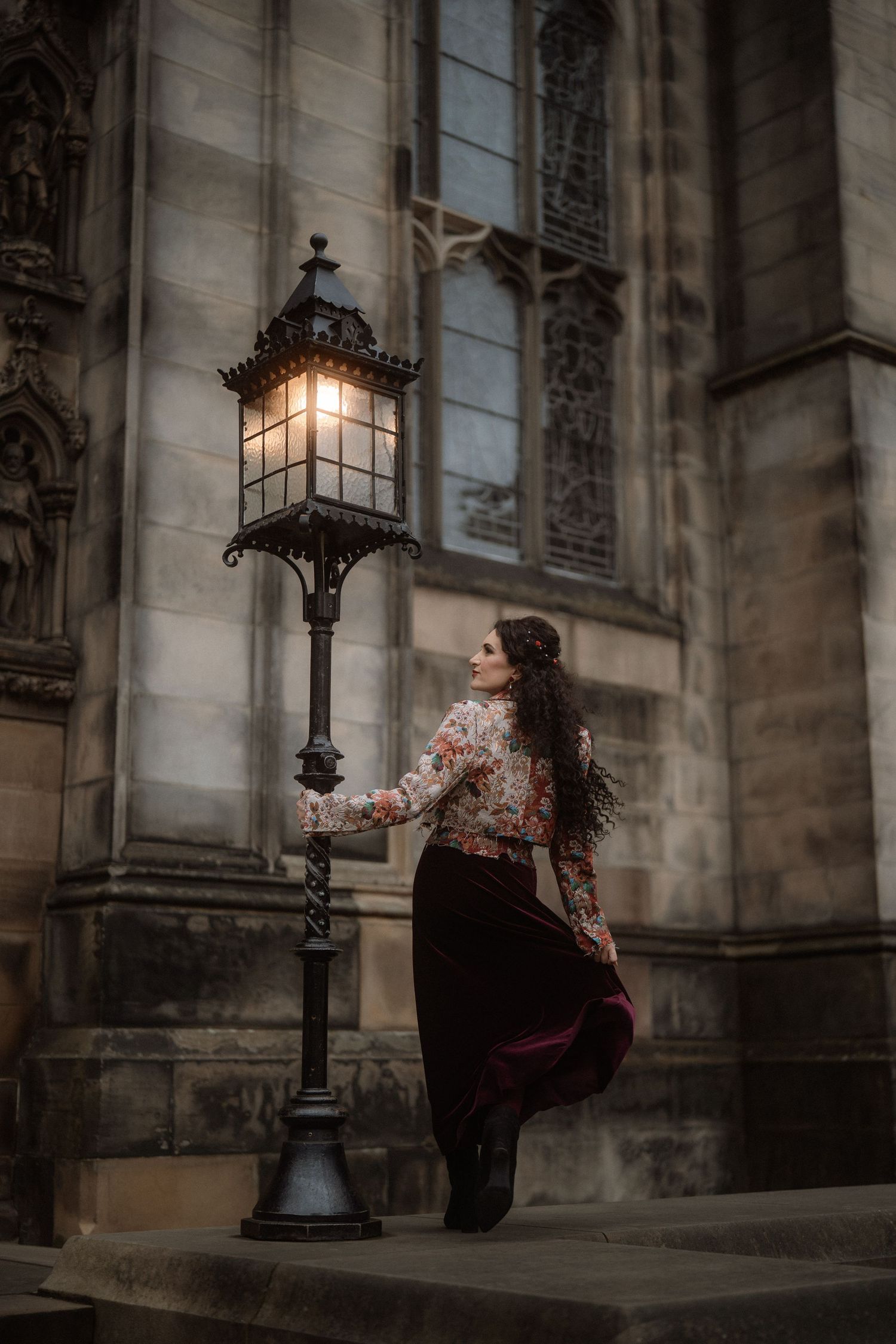 Woman in floral blouse and velvet skirt beside a glowing vintage street lamp outside a Gothic stone cathedral.