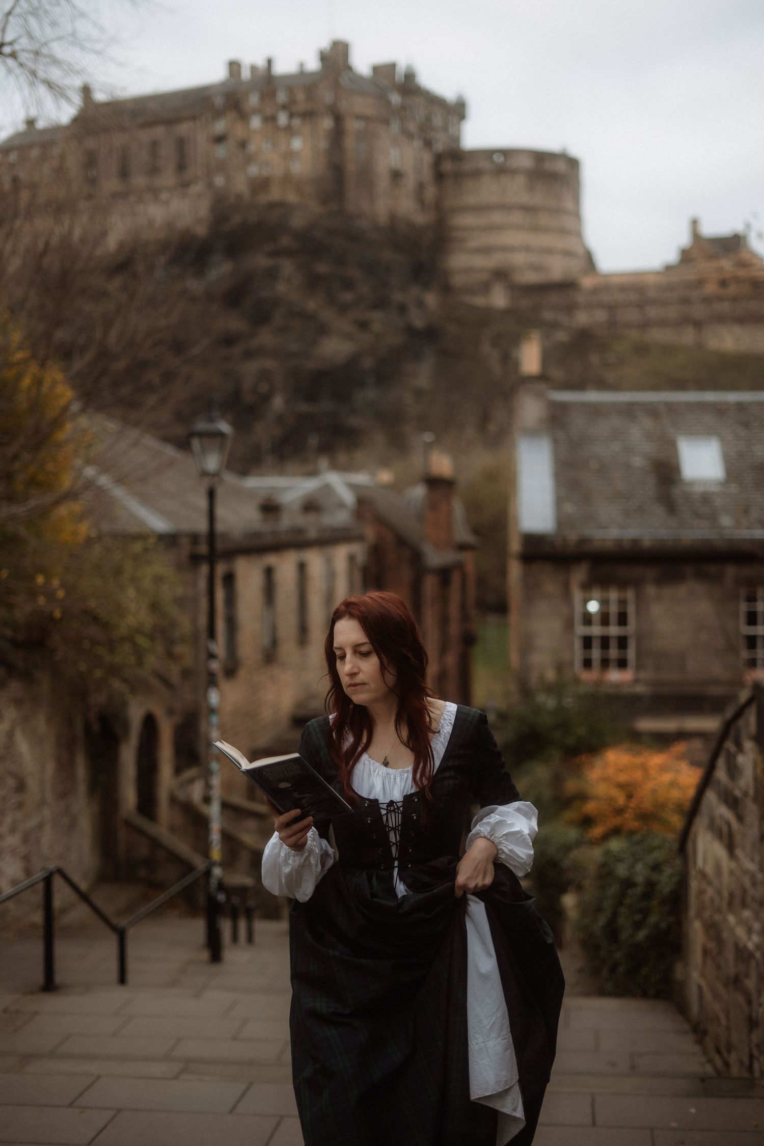 A person in period costume stands on a stone stairway with Edinburgh Castle looming dramatically in the background.