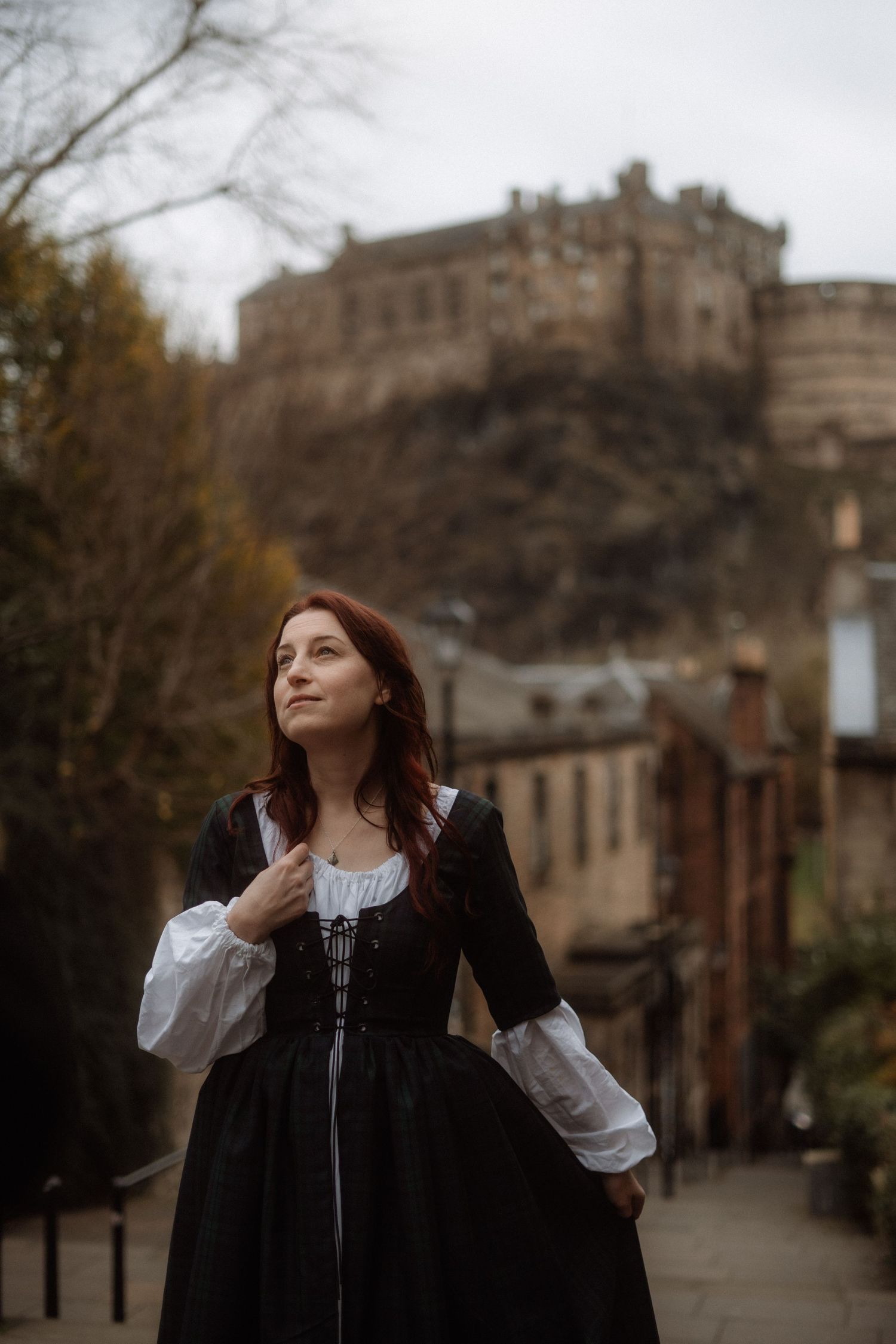 Woman in a medieval-style black dress stands on a cobblestone street with Edinburgh Castle looming in the background.