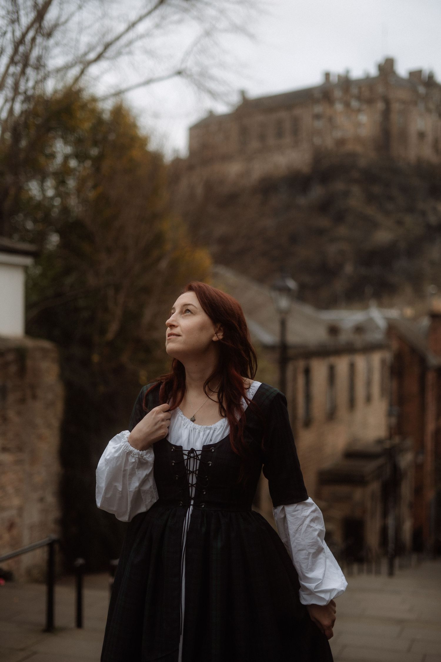 A person in a black coat and white blouse poses thoughtfully against an autumn backdrop with historic buildings.