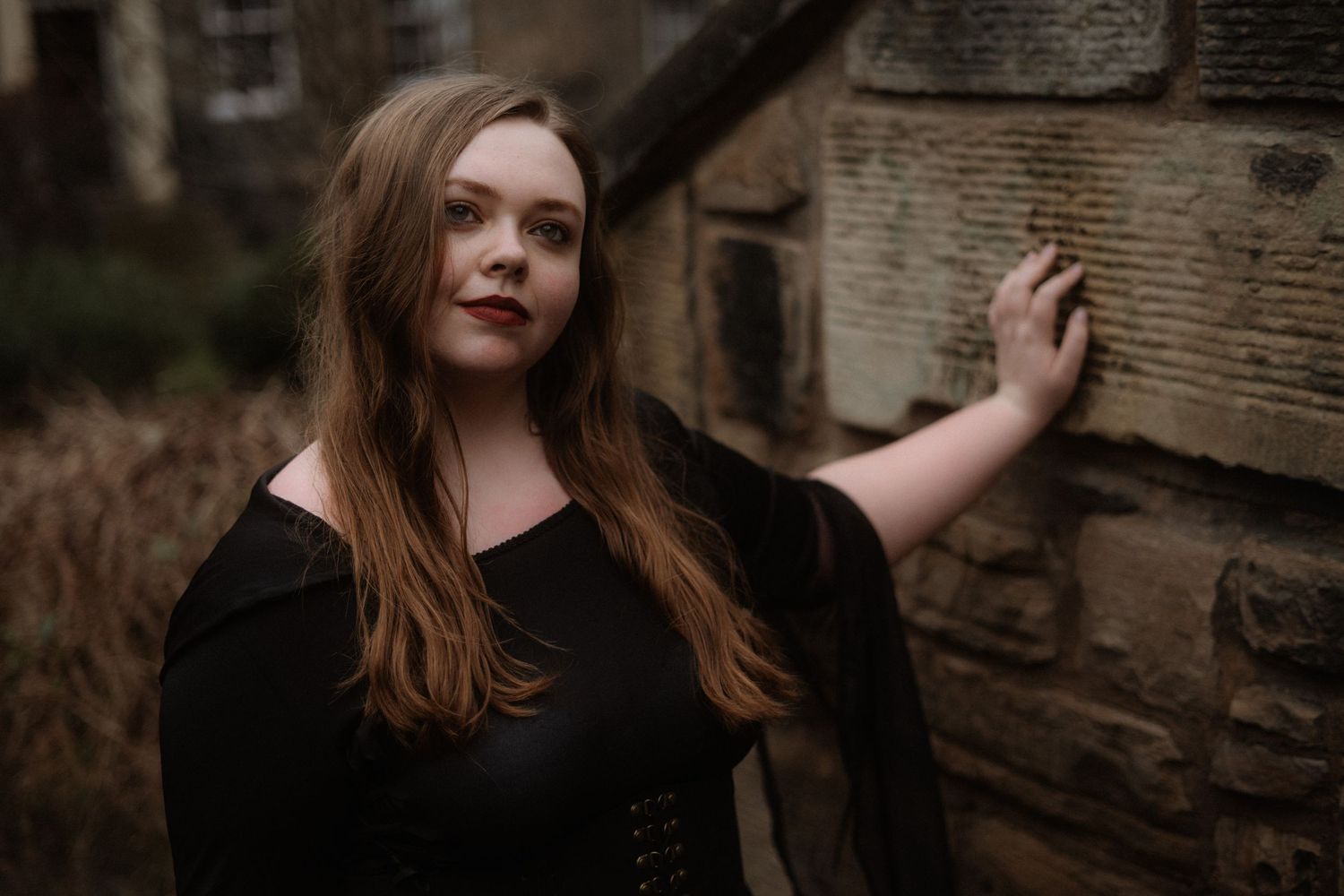 A person with long brown hair poses against a rustic wooden wall in a moody outdoor setting.