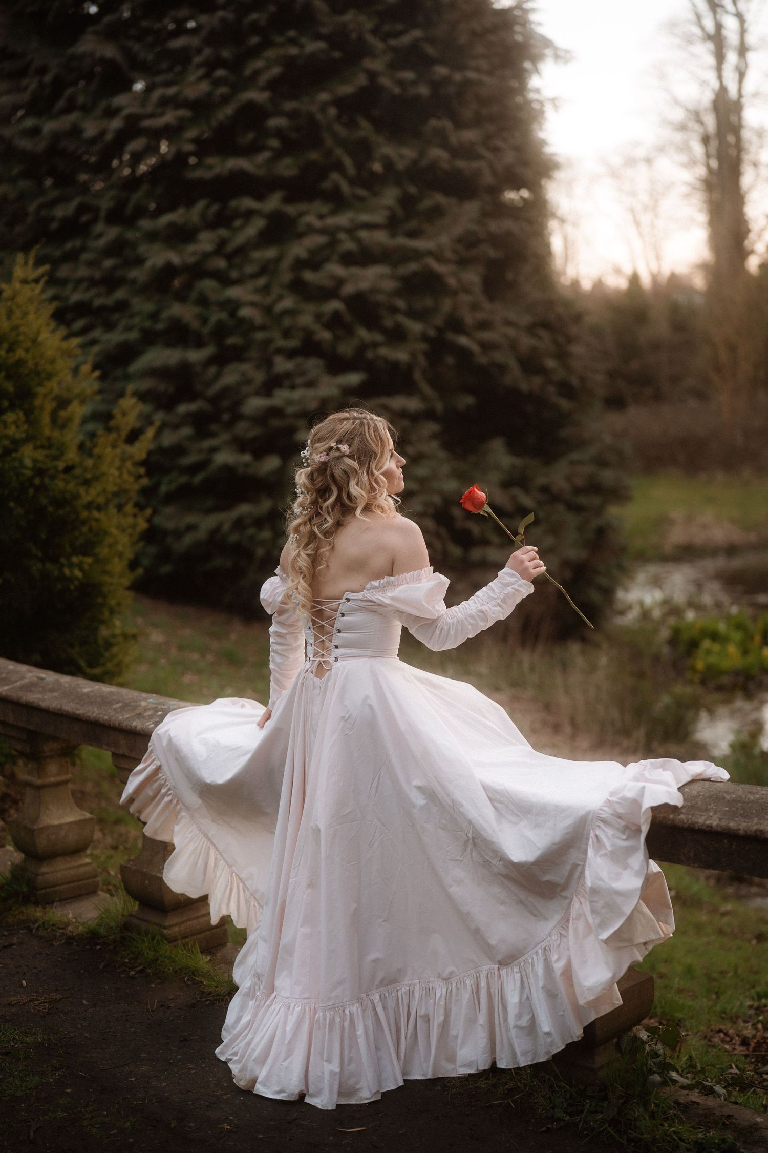Woman in white off-shoulder ballgown holding a red rose, standing in a lush garden at golden hour.