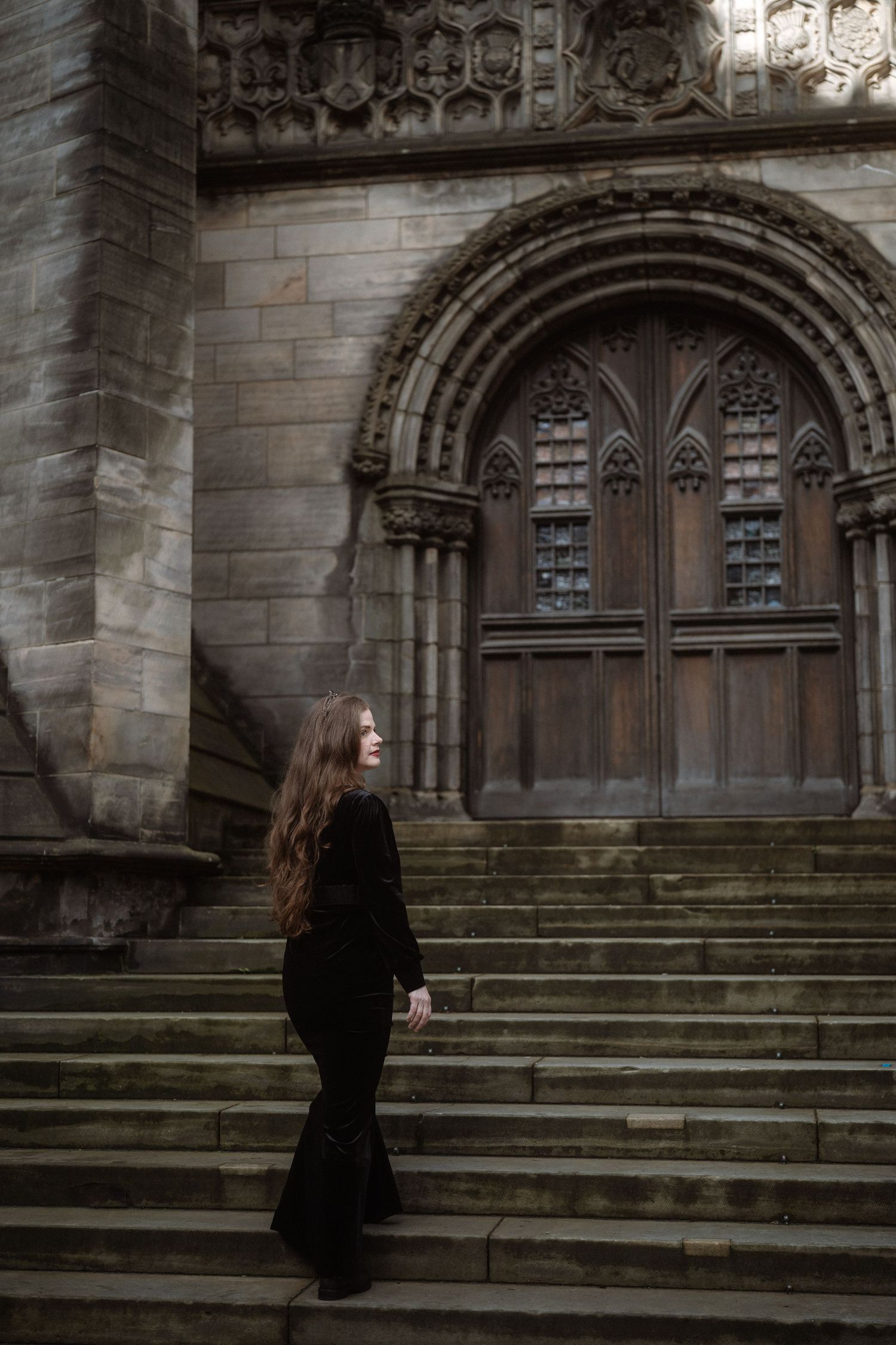 Woman in black velvet outfit standing on stone steps before a Gothic cathedral's ornate arched wooden door.