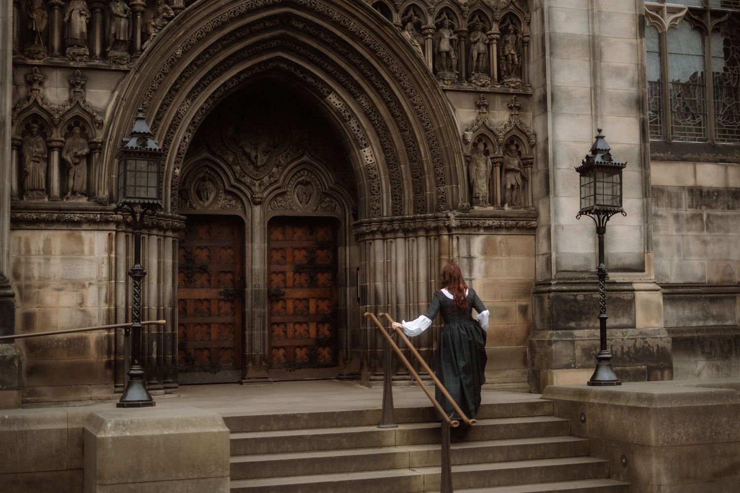 Gothic cathedral entrance with ornate stone archways and wooden doors illuminated by vintage lampposts in evening light.