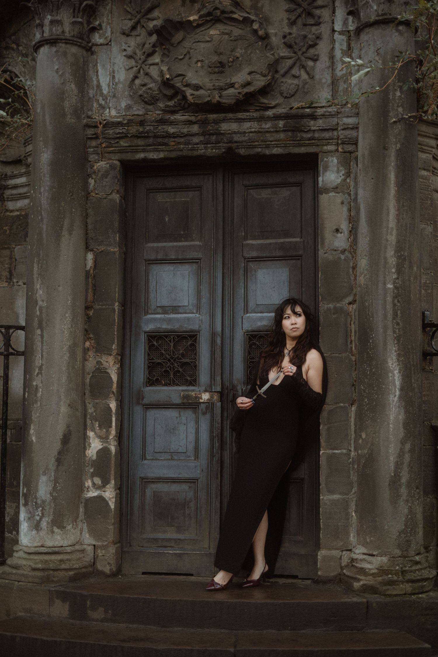 A person in black dress sits in an old stone doorway with ornate architectural details and weathered blue doors.