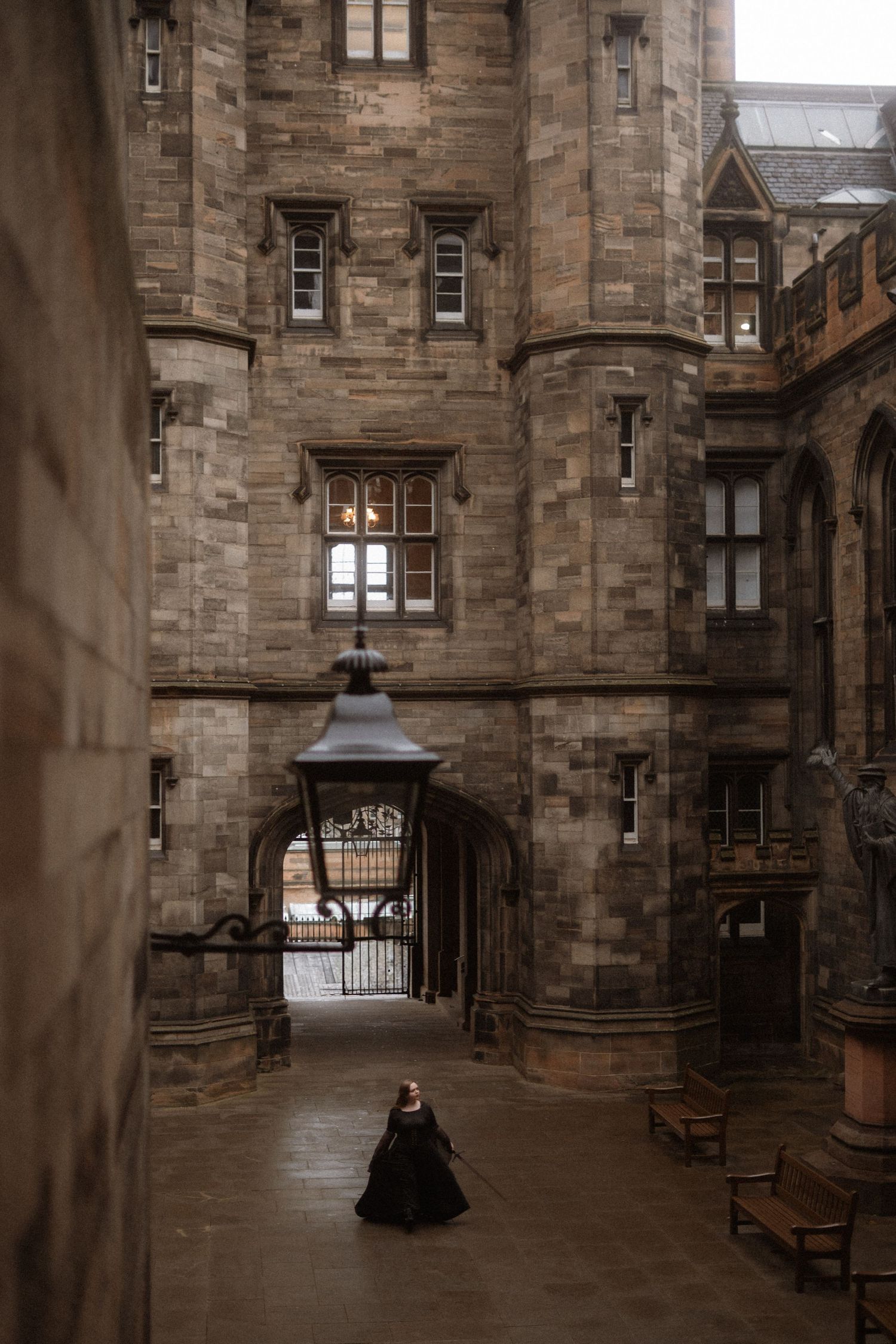 A figure in a black dress stands alone in a gothic stone archway corridor at a historic university building.
