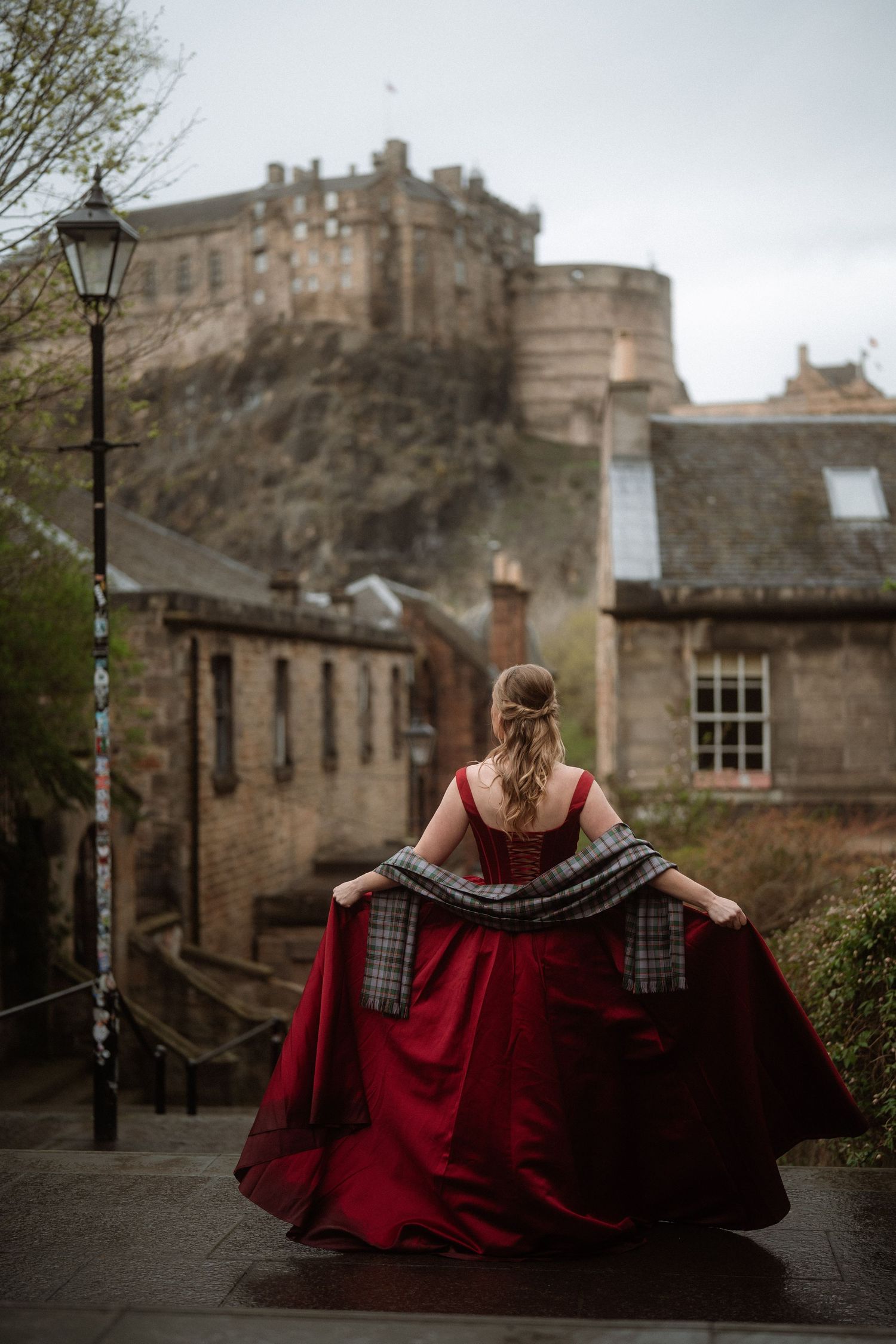Woman in red gown with tartan shawl on Edinburgh Old Town steps, Edinburgh Castle visible in background.