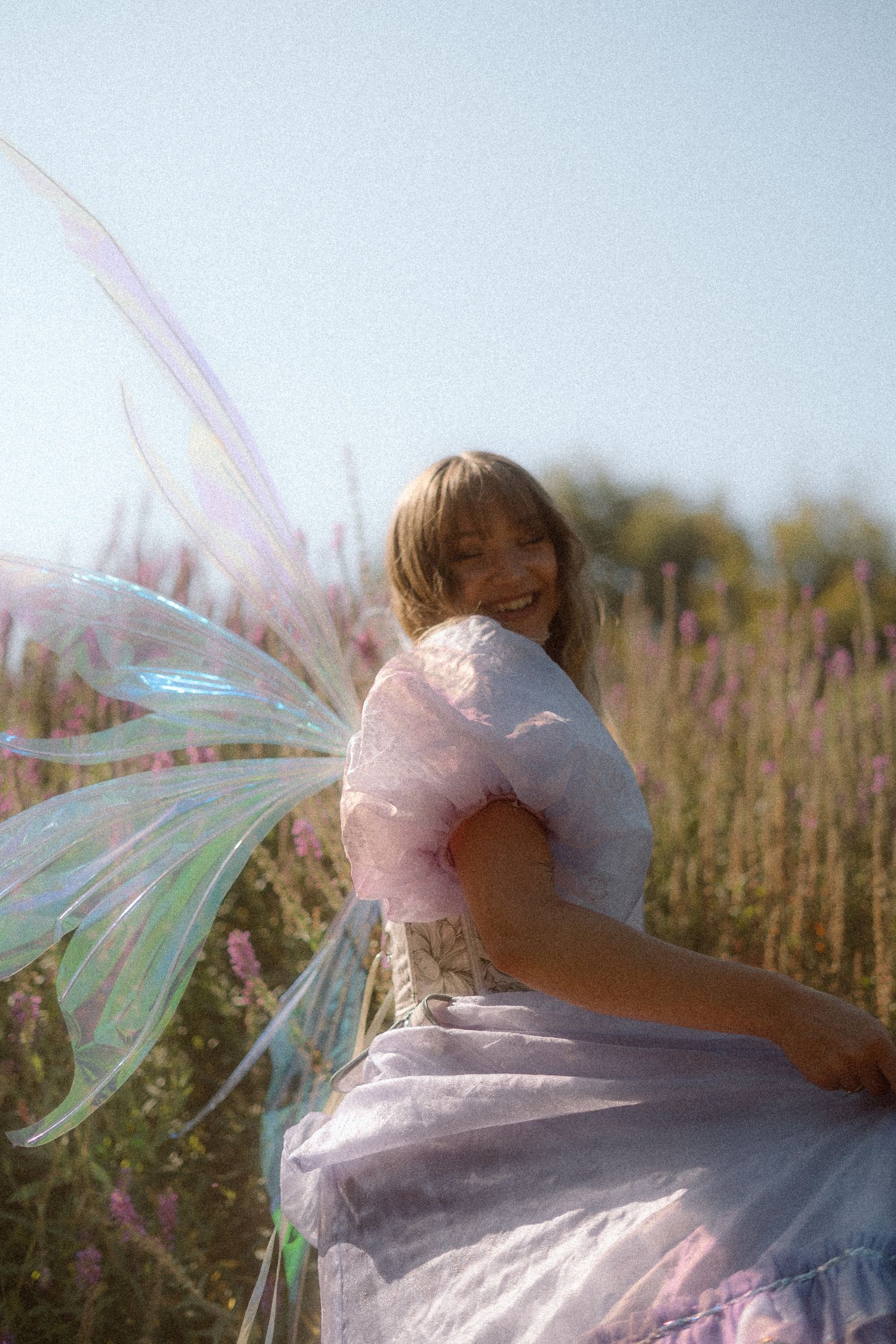 A person in a dreamy fairy costume with iridescent wings poses playfully in a sunlit meadow with wildflowers.