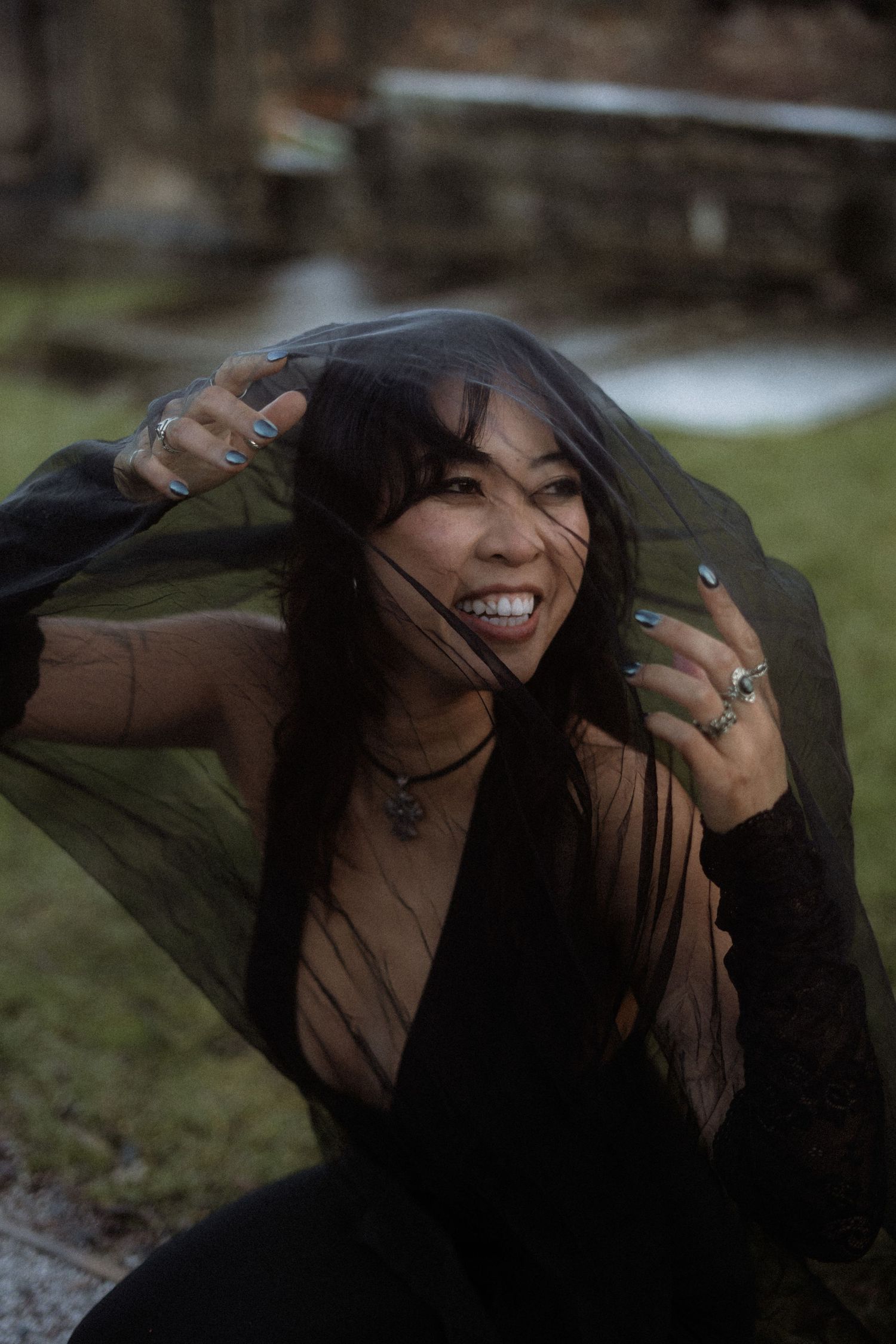A person playfully hides beneath a sheer black fabric veil while smiling outdoors in natural lighting.