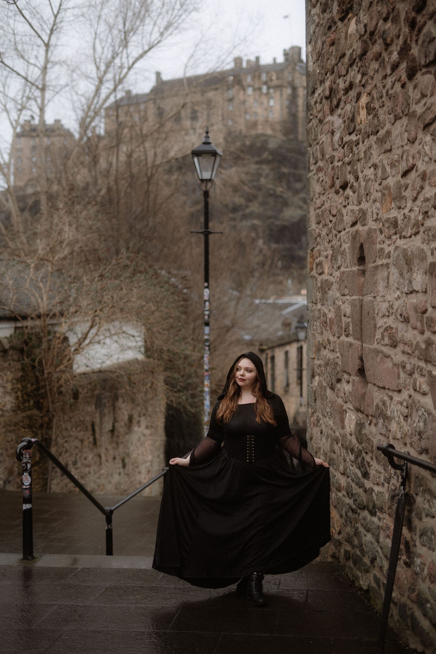 A figure in a flowing black dress poses dramatically against stone castle walls on a moody winter day.