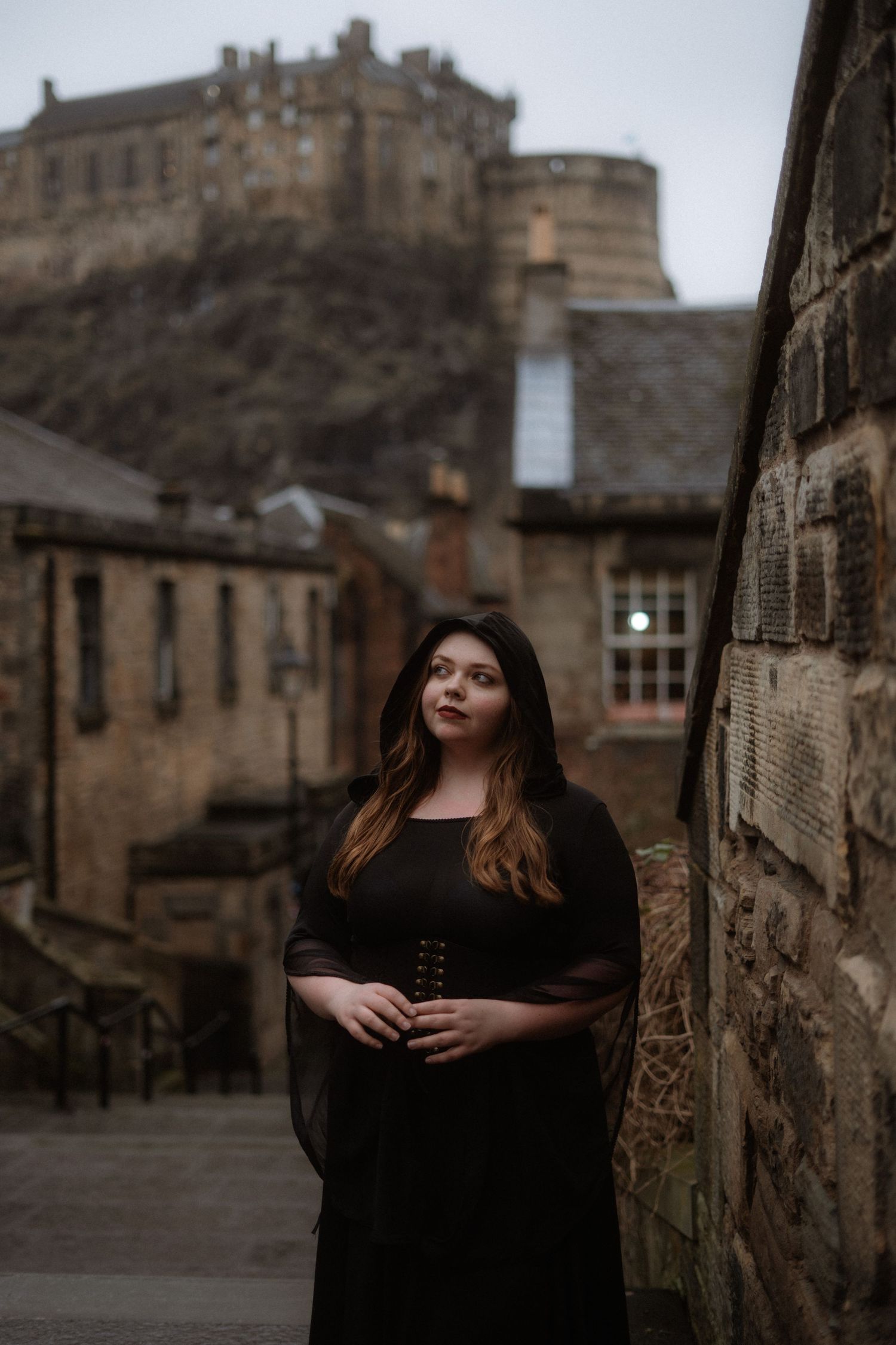 A figure in a black dress poses dramatically against historic stone buildings with Edinburgh Castle visible in the background.