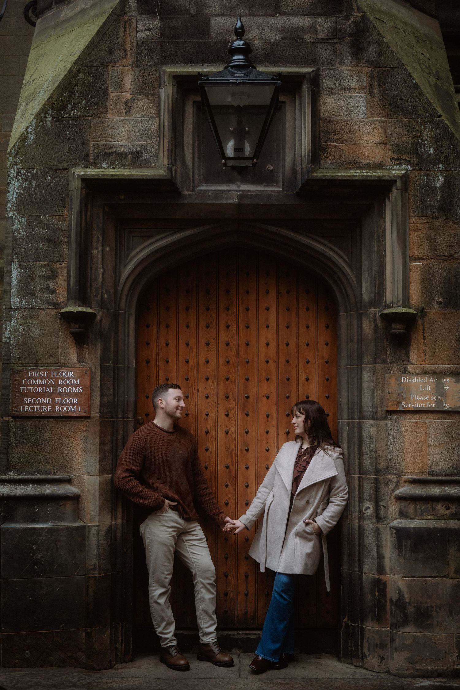 Couple holding hands in front of a grand wooden arched door at a historic stone building.
