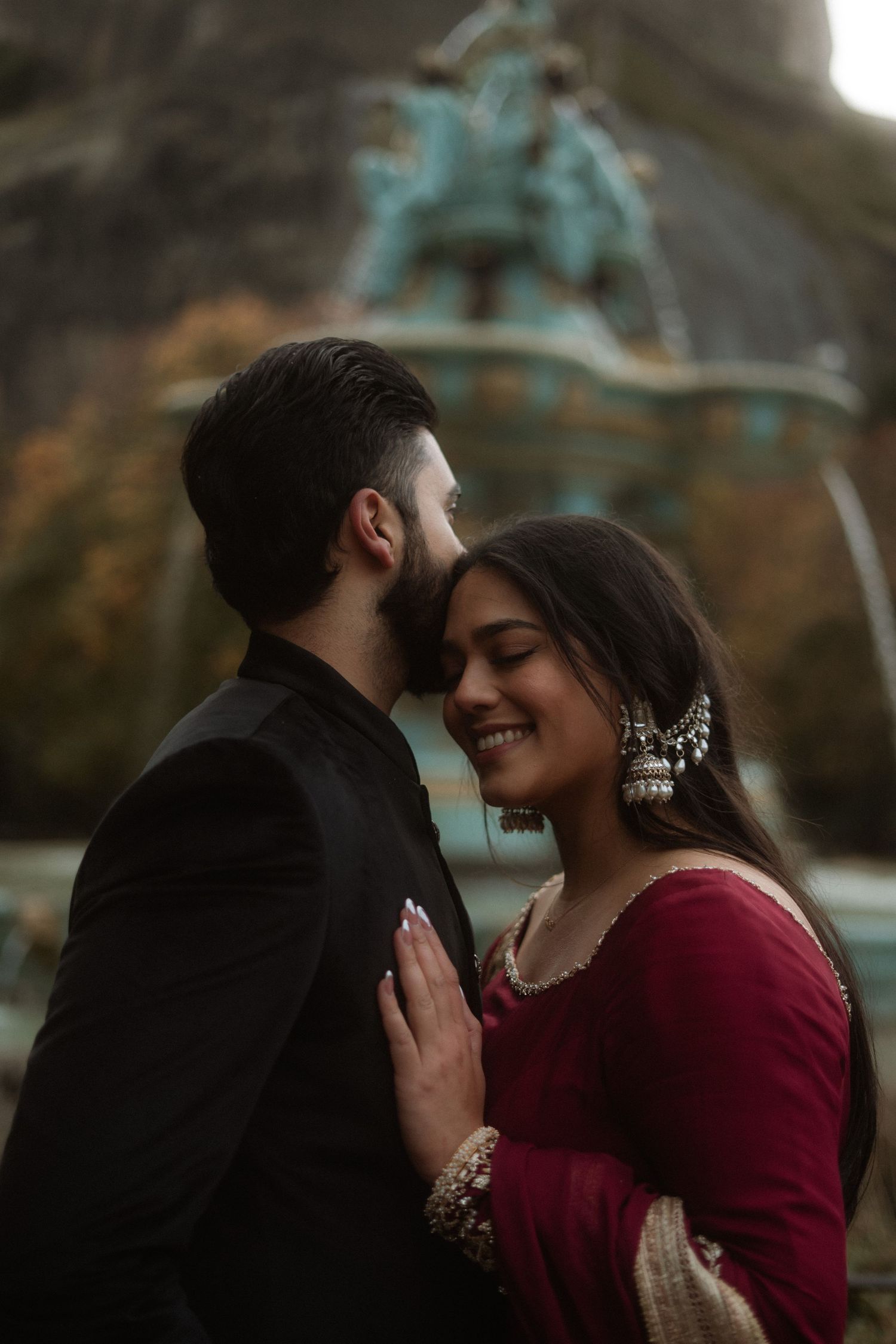 Couple in traditional South Asian attire sharing a romantic moment near a fountain statue outdoors.
