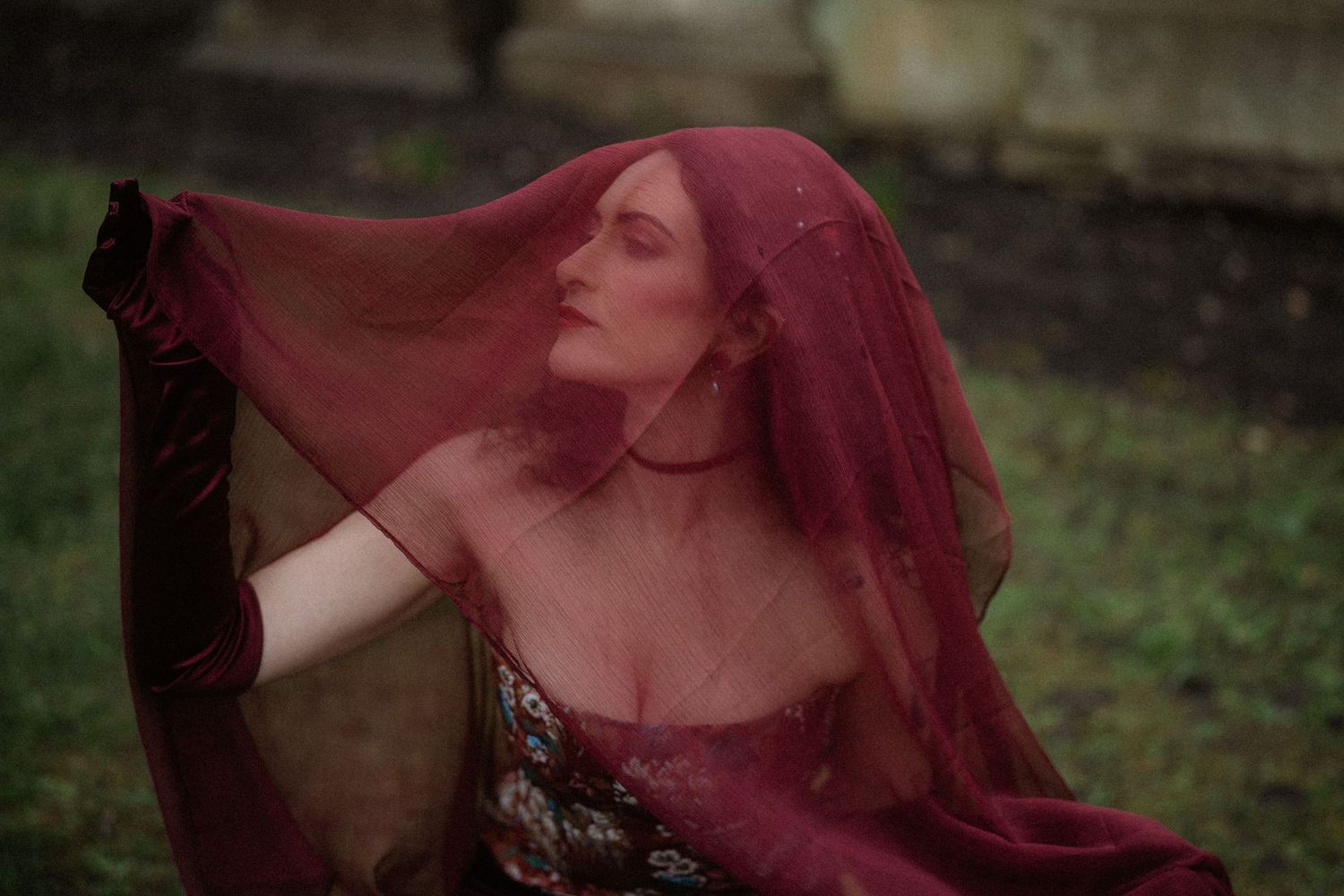 A woman in a dark lace corset holds a flowing burgundy veil outdoors in a moody, gothic-inspired portrait.