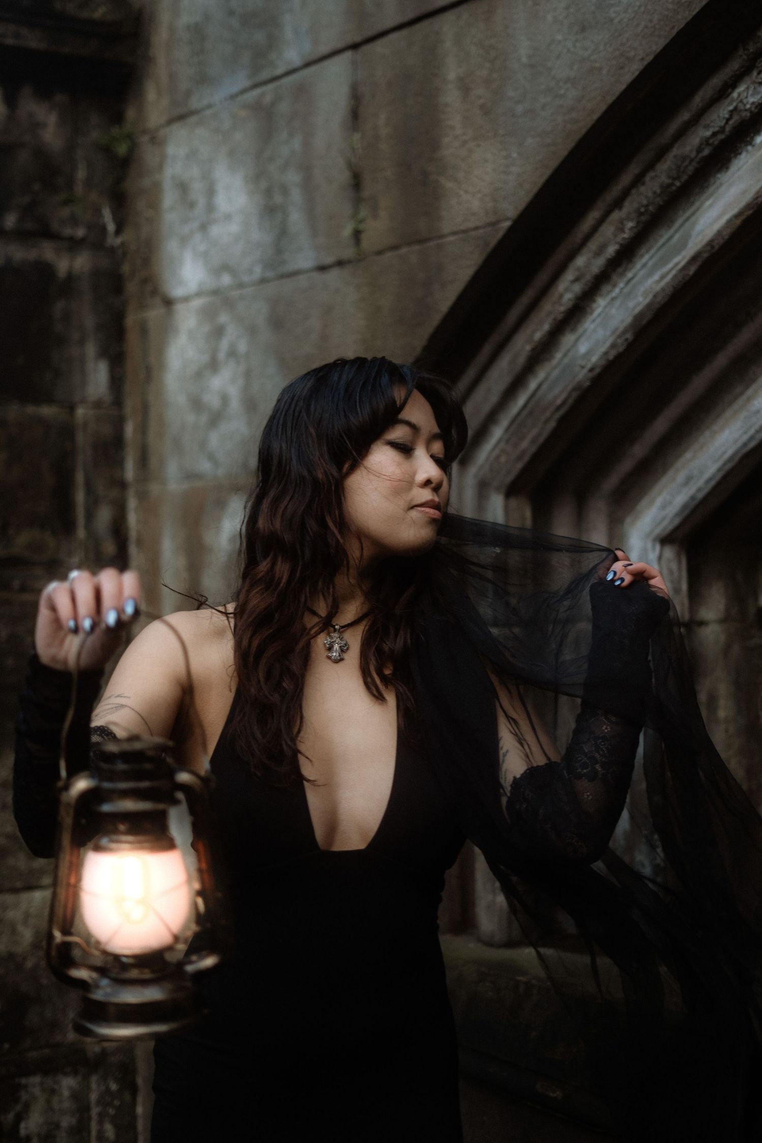 A person in dark clothing holds an antique lantern while exploring a dimly lit, atmospheric space.