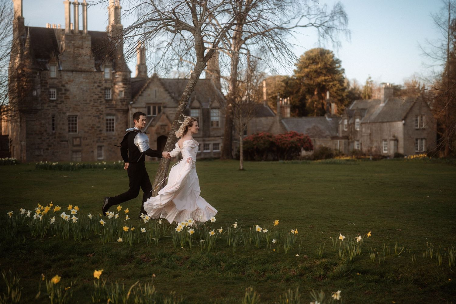 Bride and groom running through spring daffodils on lush green lawn before a grand historic stone manor house.