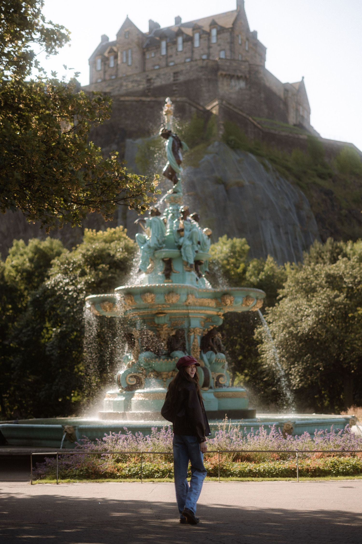 Person standing before the ornate Ross Fountain in Princes Street Gardens with Edinburgh Castle on volcanic rock behind.