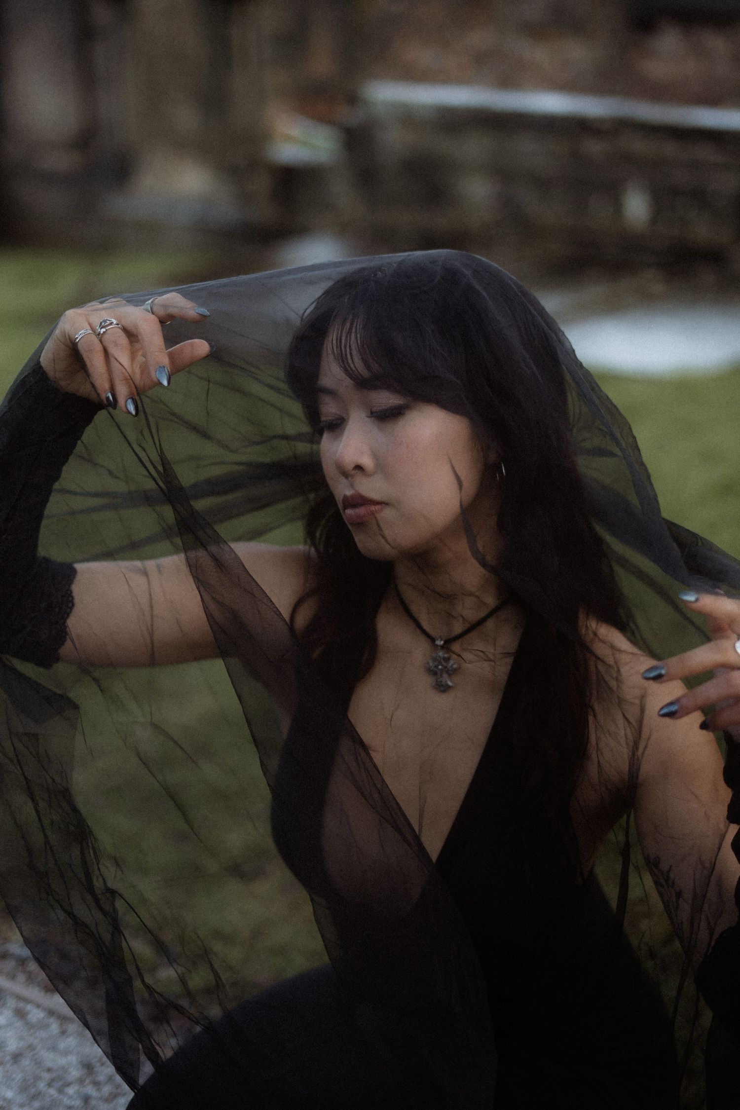 A person in dark clothing poses with a black veil against a blurred outdoor background in a moody photoshoot.