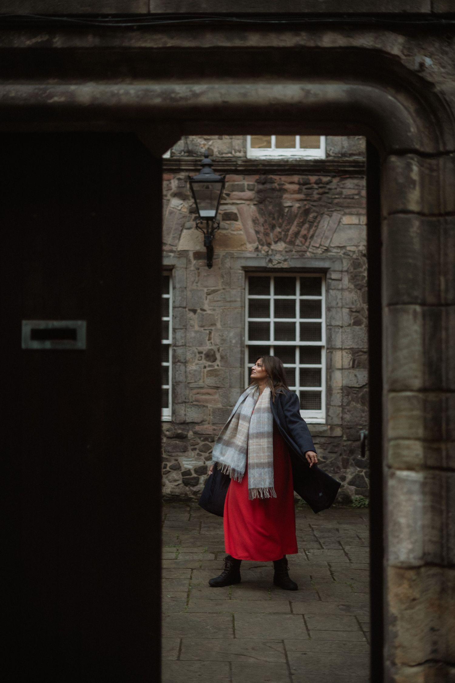 Woman in red skirt and plaid scarf standing in historic stone archway courtyard.