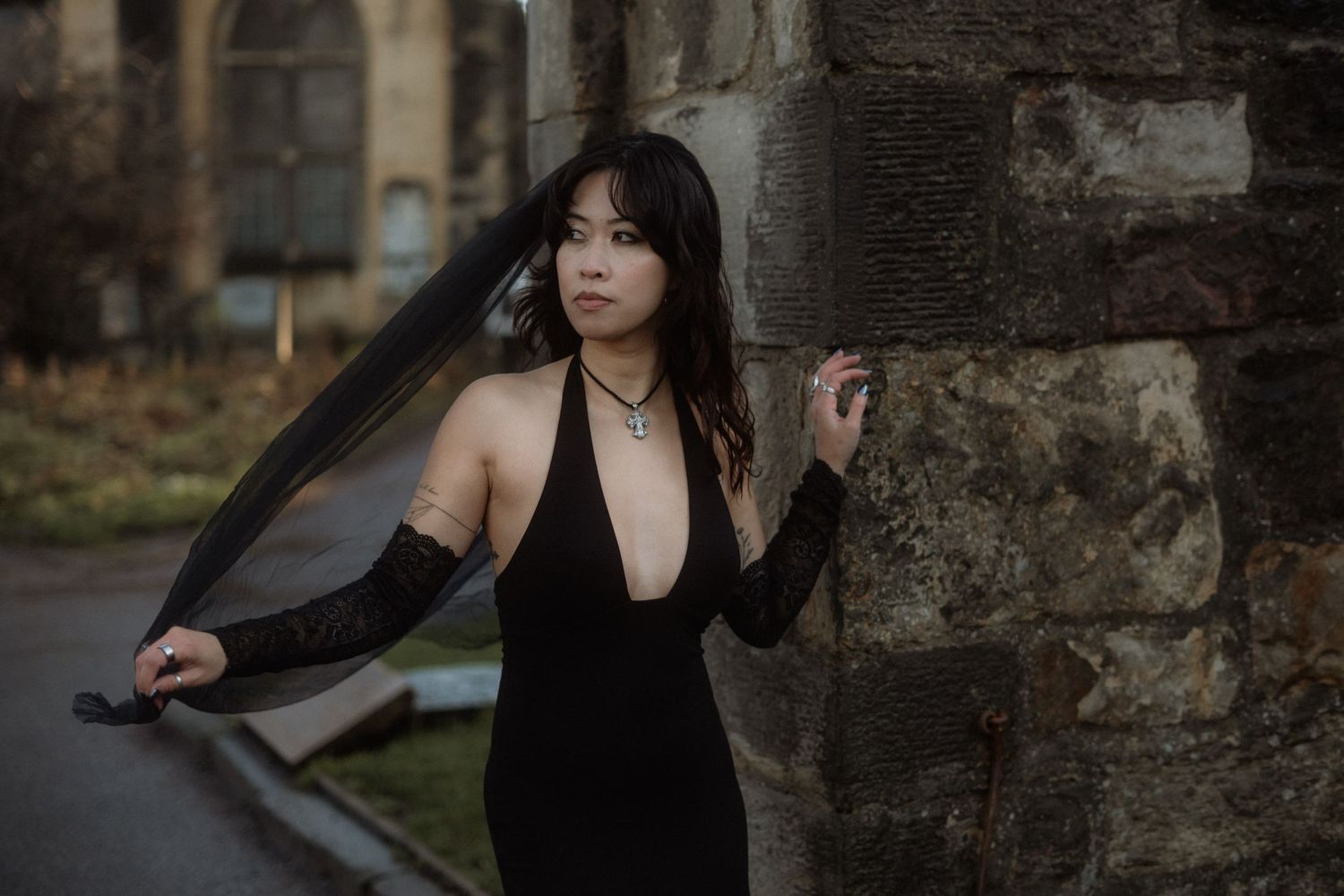 A figure in an elegant black dress poses against a stone wall while holding sheer fabric flowing in the wind.