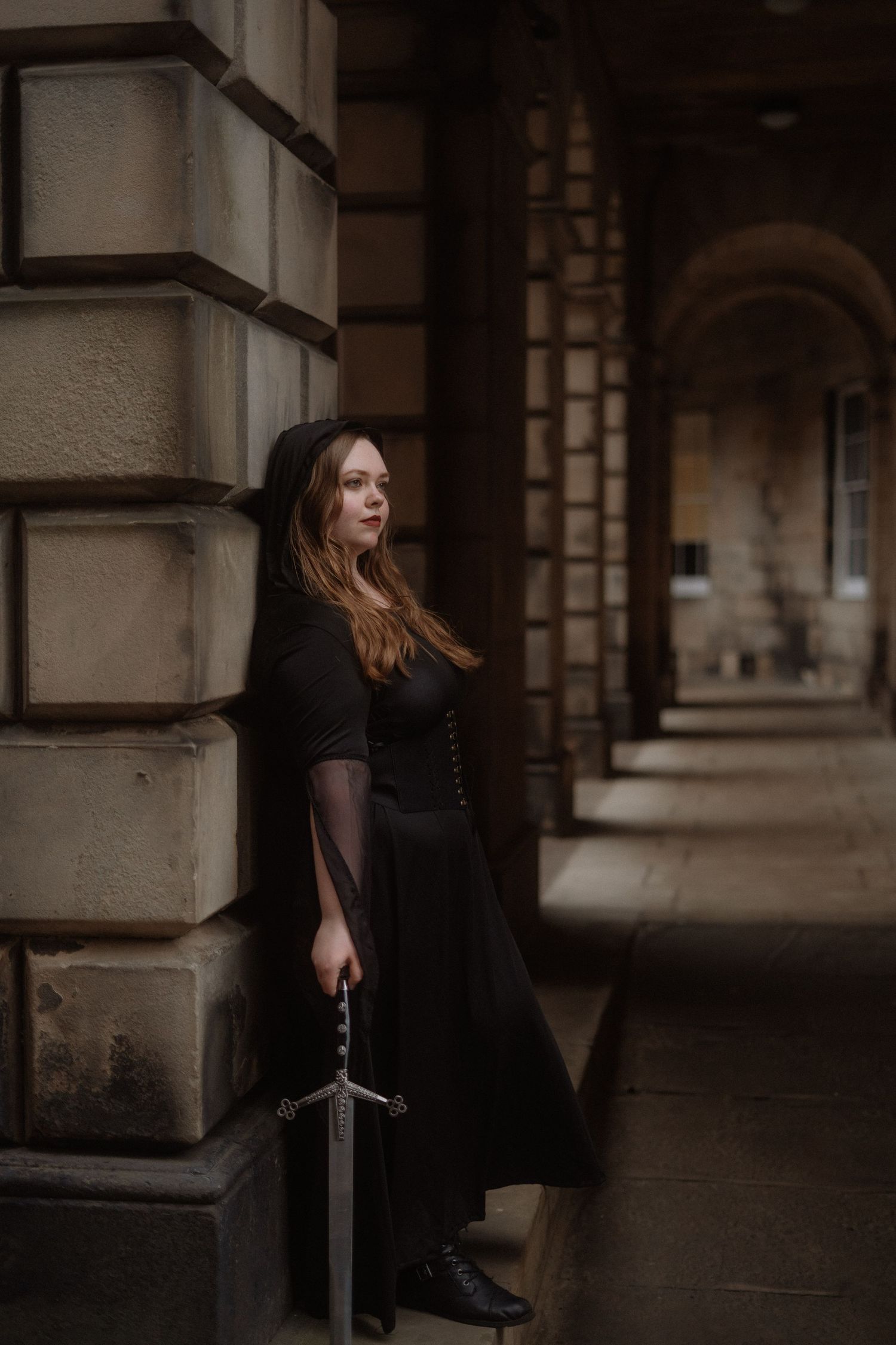 A person in a black dress stands in a moody architectural setting with stone columns and arches.