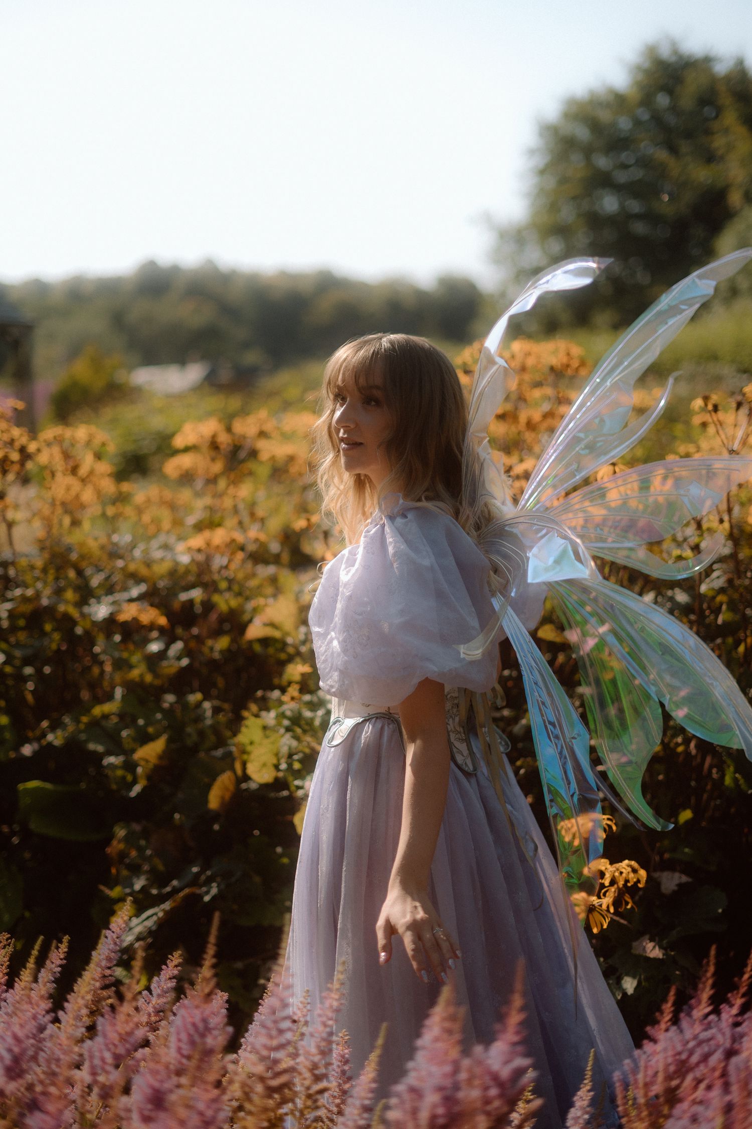 A figure in a flowing white dress stands among pink flowers and sunflowers in a dreamy garden setting.