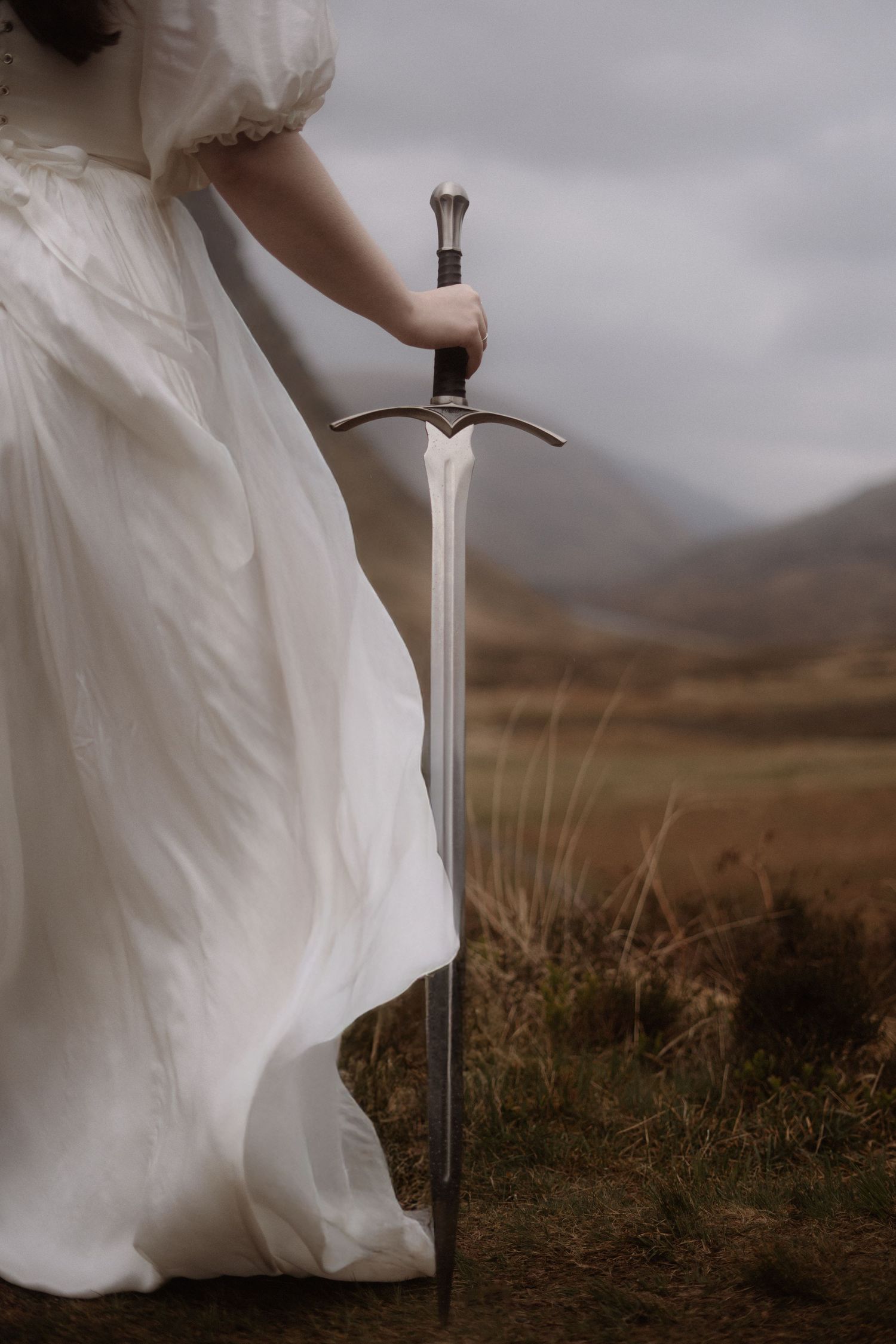 A medieval sword stands upright in a dramatic landscape with flowing white fabric against a moody sky in Glencoe, Scotland.