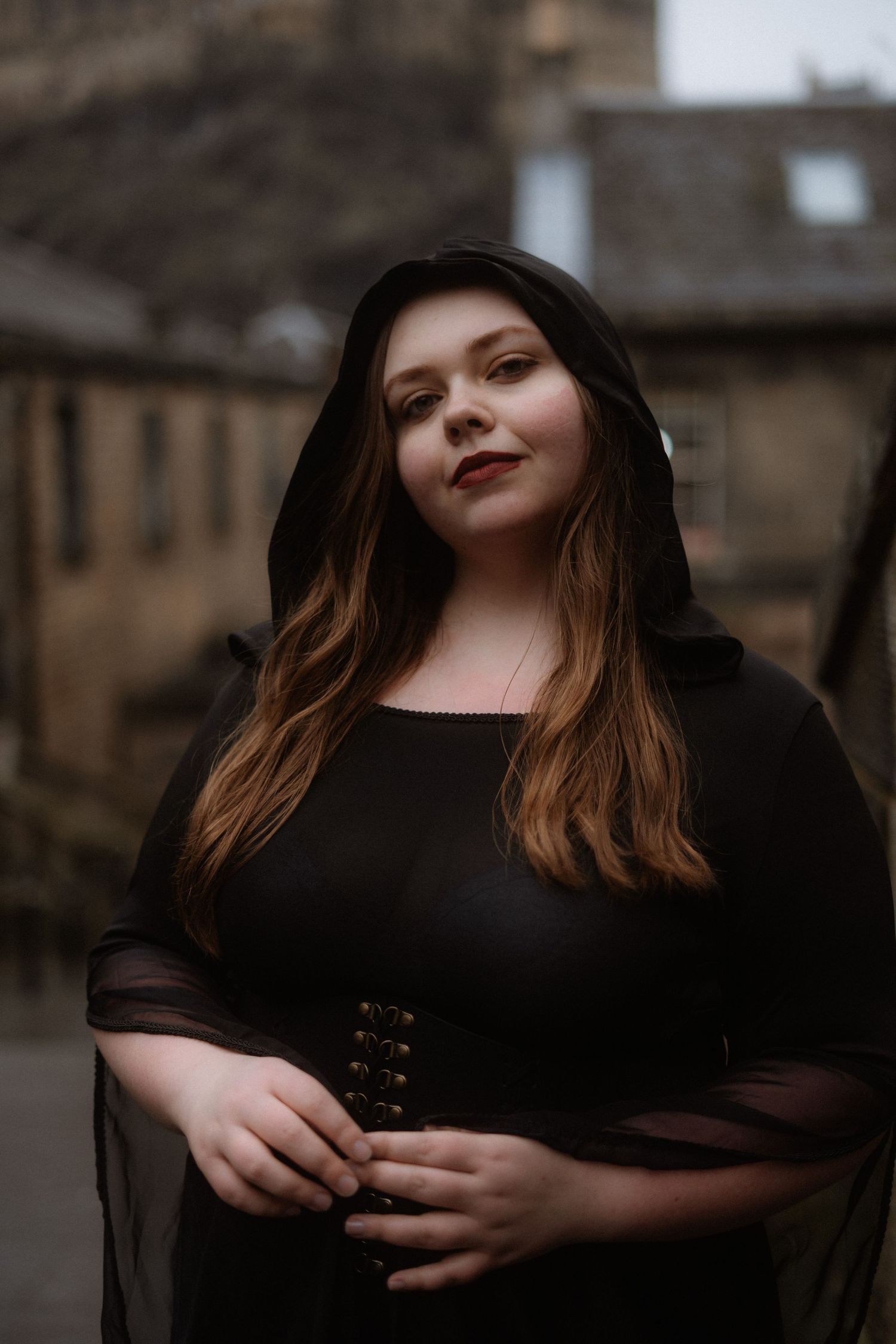 A person wearing a black dress with sheer sleeves and a hood poses outdoors against a moody architectural backdrop.