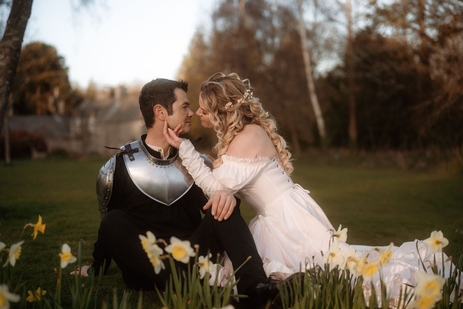 A couple in medieval wedding attire share a romantic kiss among spring daffodils, groom in armor and bride in white gown.