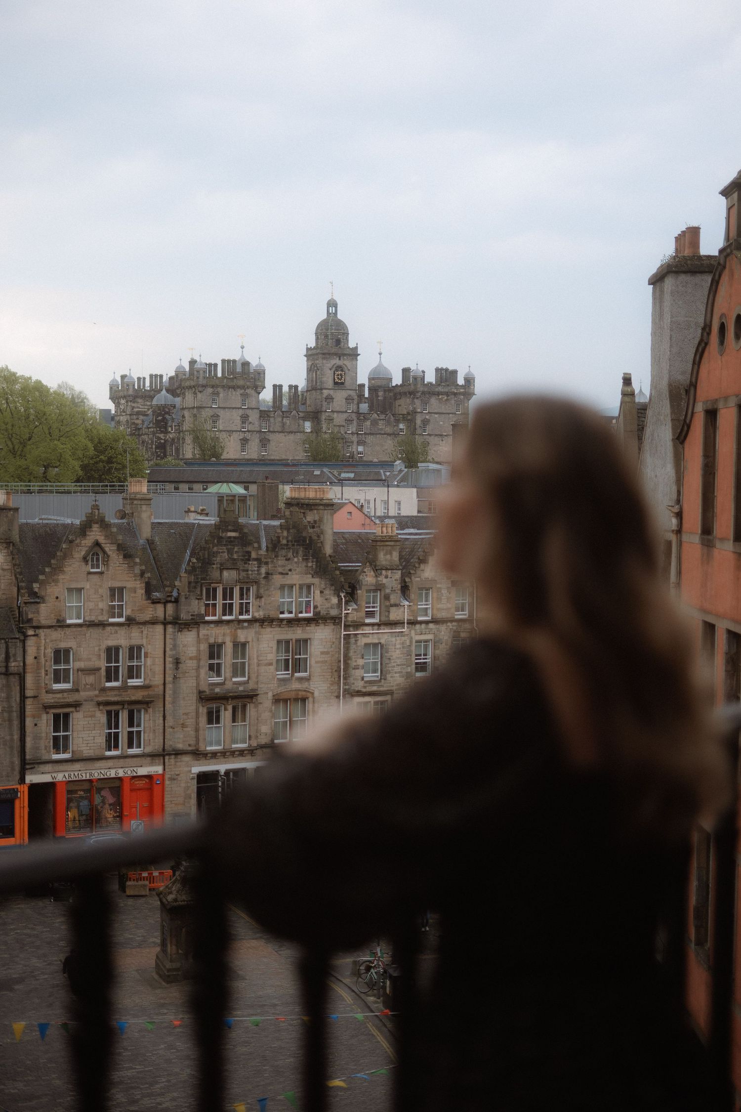Woman gazing at historic Edinburgh stone buildings and castle from a balcony railing on a cloudy day.
