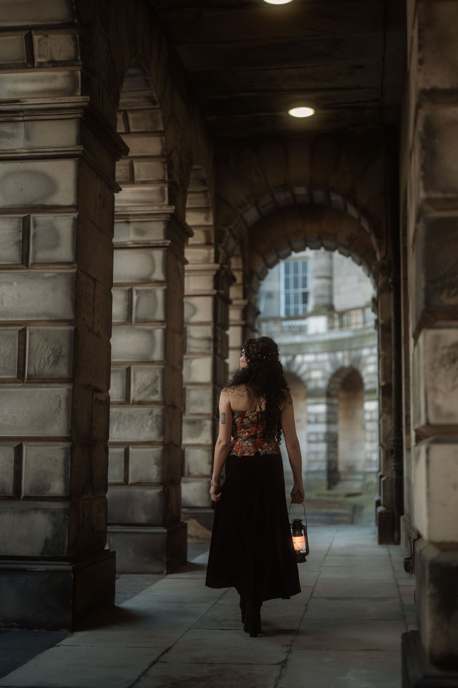 Woman in floral corset and black skirt walks through stone archway corridor holding a vintage lantern.