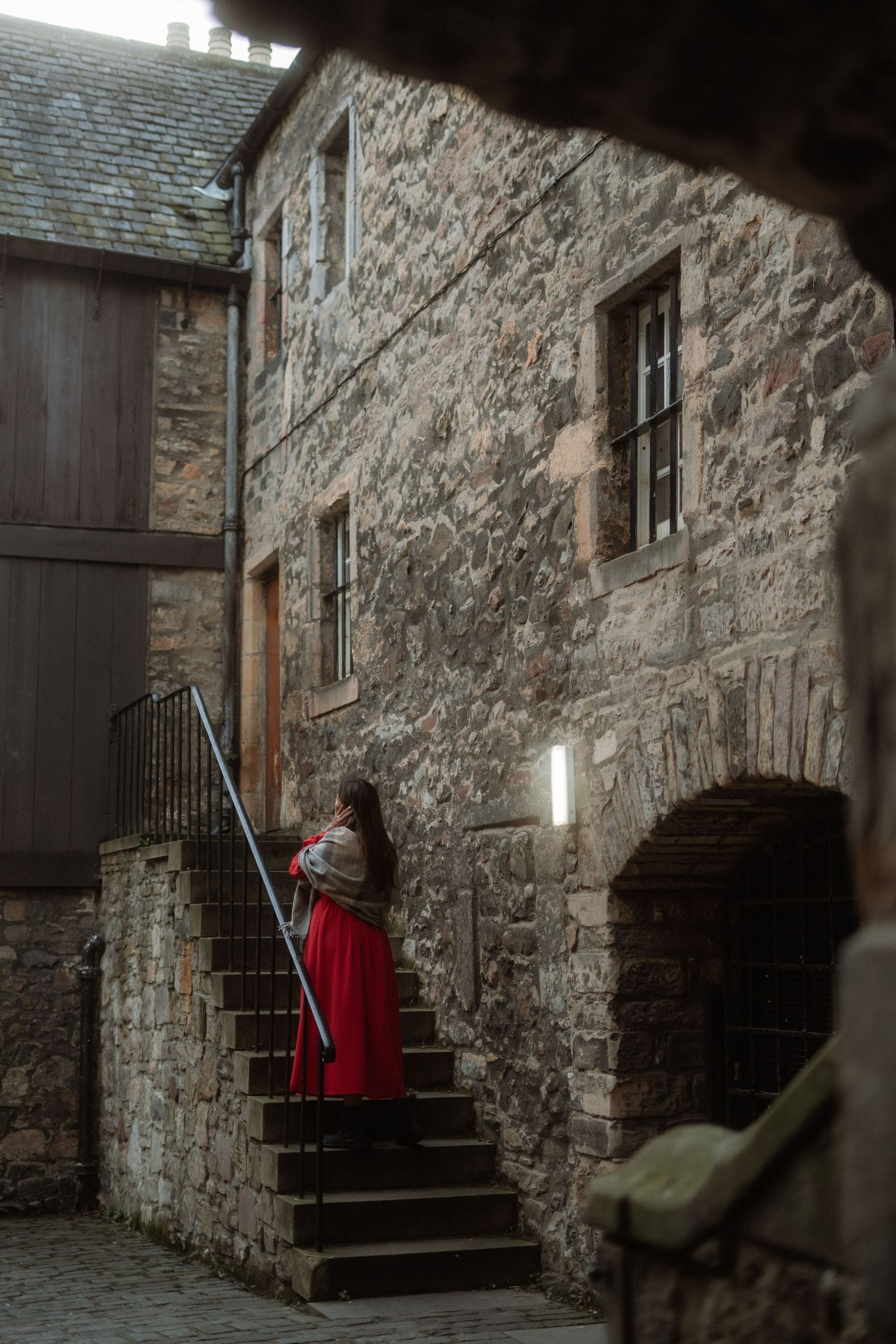 Woman in red dress standing on stone steps beside historic medieval stone building with warm wall lighting.
